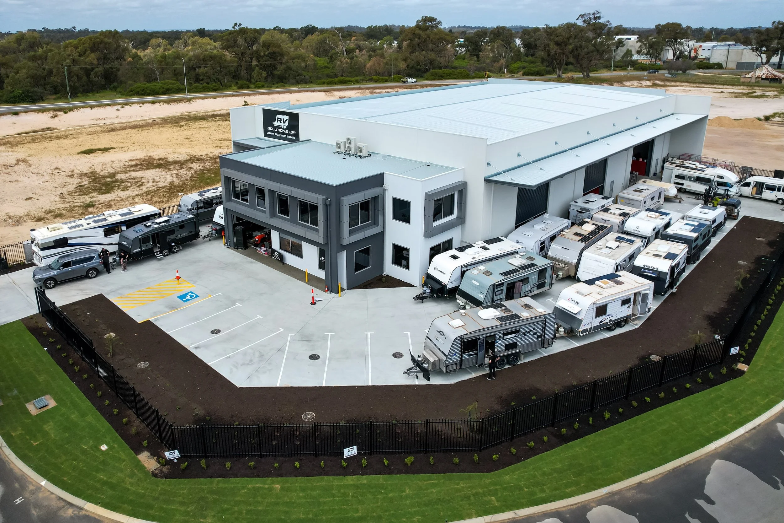 Aerial view of a business building with RV dealership and storage, surrounded by parking lot with RVs and cars, fenced yard, and green lawn.