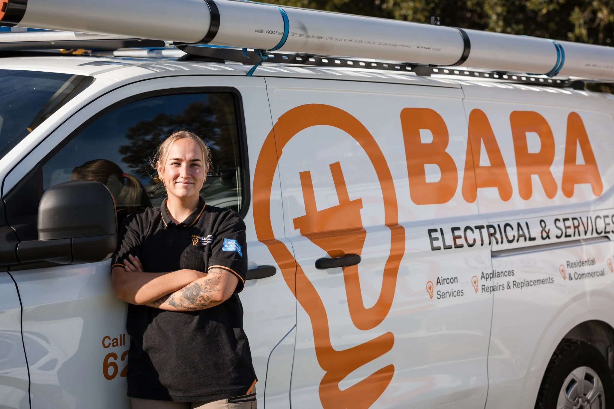 Woman standing with arms crossed in front of a white van with orange and black branding for BARA Electrical & Services, equipped with a ladder and pipes on the roof.