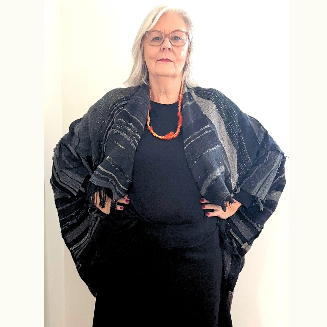 Grey haired woman with glasses dressed in all black wearing a black handwoven jacket stands with her hands on her hips