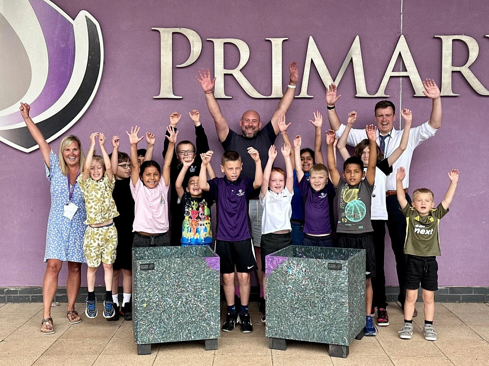 Eco Leaders and teachers form Nottingham Primary Academy smiling and raising their hands in front of the school and the two recycled plastic planters they made