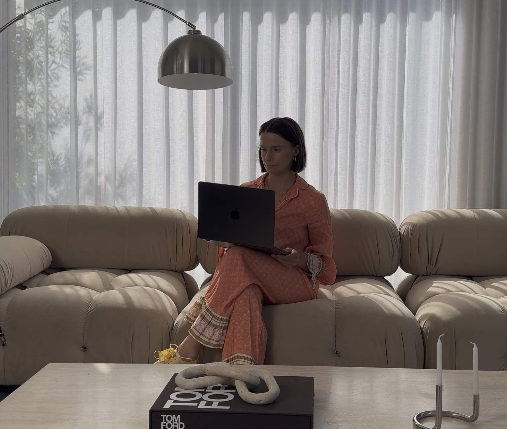 A woman sitting on a light-colored sofa in front of sheer curtains, working on a laptop. In front of her is a coffee table with a book and decorative items, including a chain and two candles.