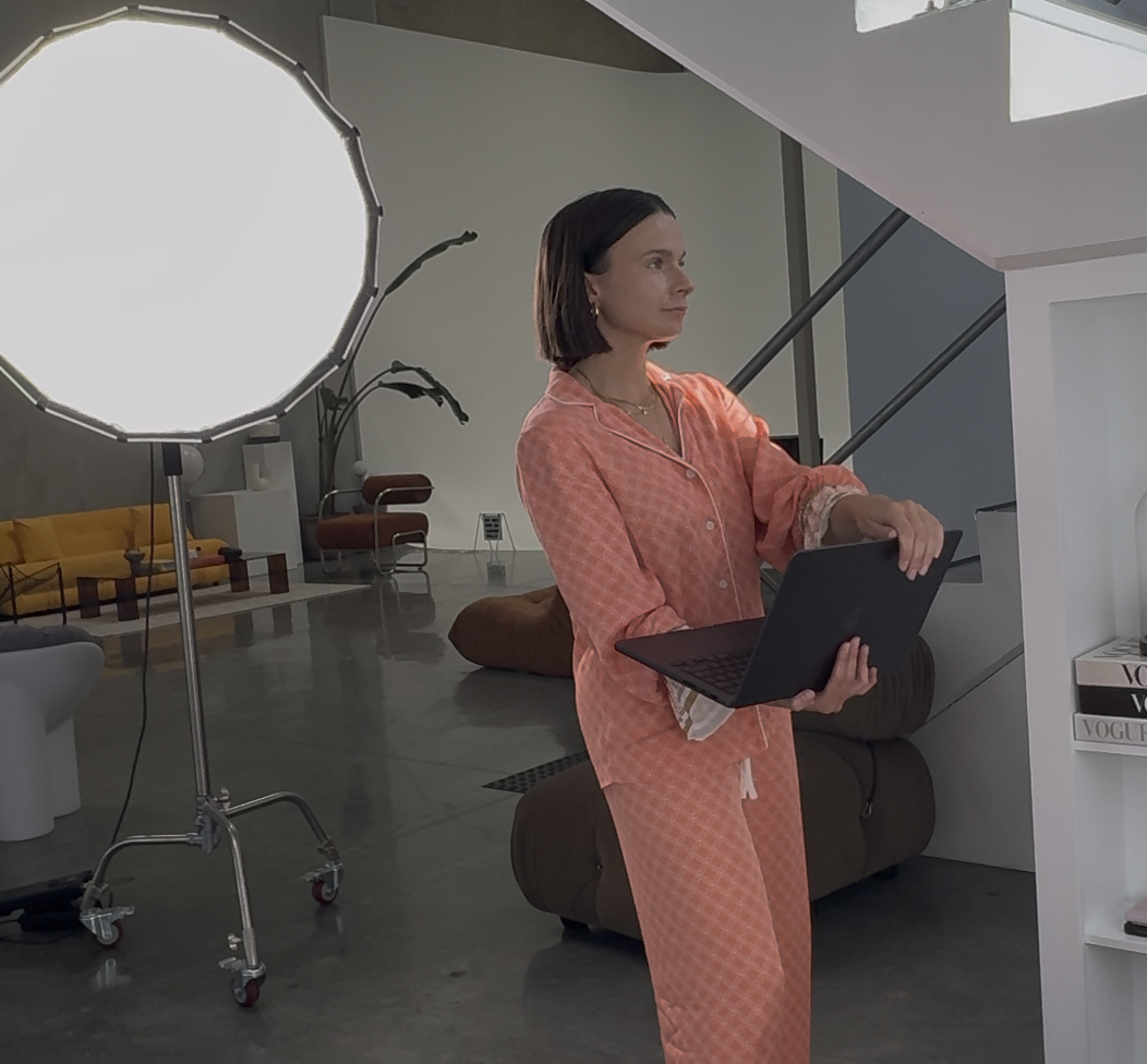 A woman wearing pink pajamas holding a laptop during a photoshoot in a modern living room with a large softbox light on a stand.