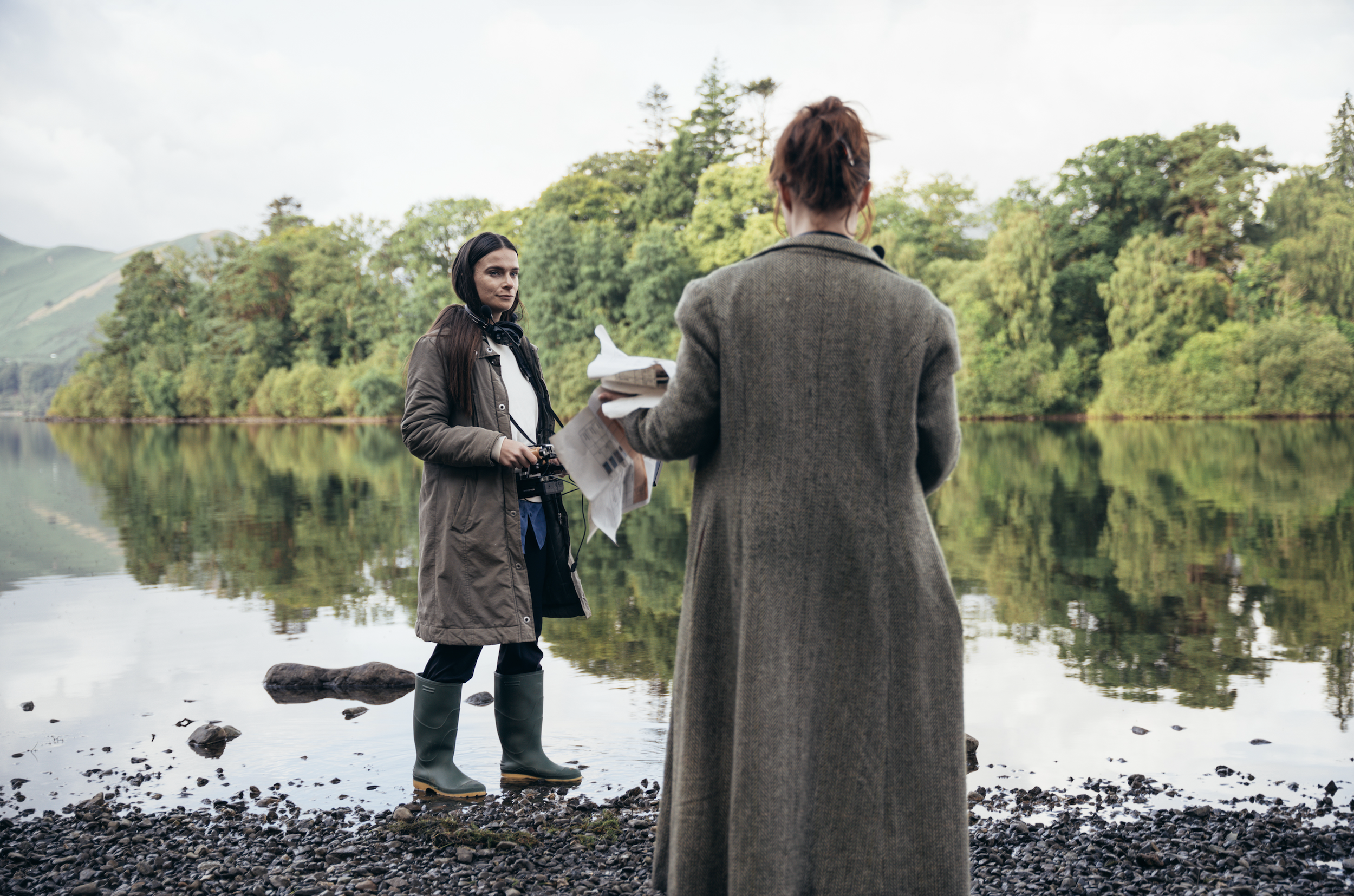 Two women talking near a lake with green trees and mountains in the background. One woman is facing away, holding papers, and the other woman is facing toward her, wearing rubber boots and holding a camera.