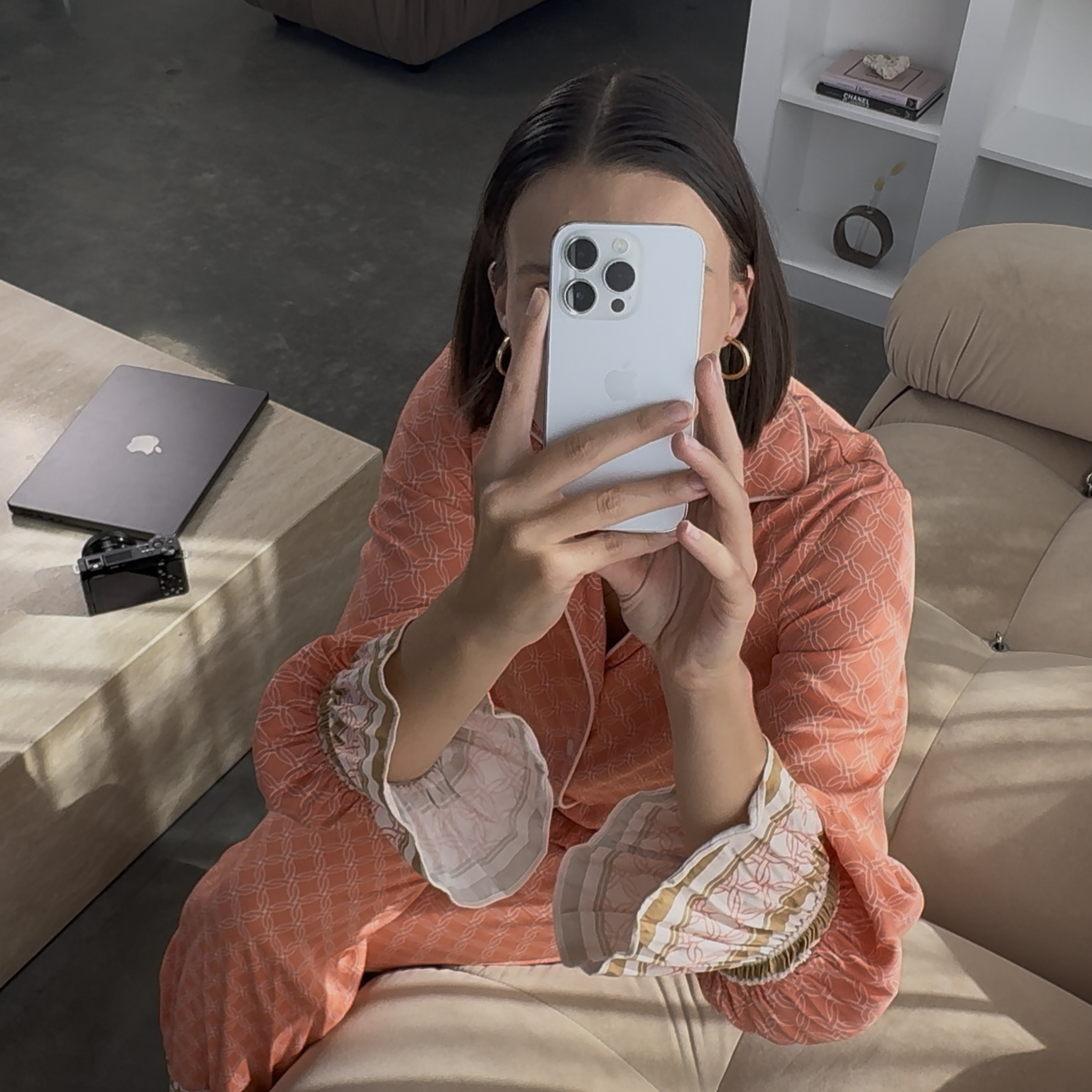 A woman with shoulder-length dark hair, wearing earrings and a patterned, coral-colored dress with light-colored, striped cuffs, is taking a mirror selfie with a white iPhone. The setting appears to be a modern living room with a beige couch, a wooden coffee table, a closed MacBook, an air camera, and shelves with books and decorative items.