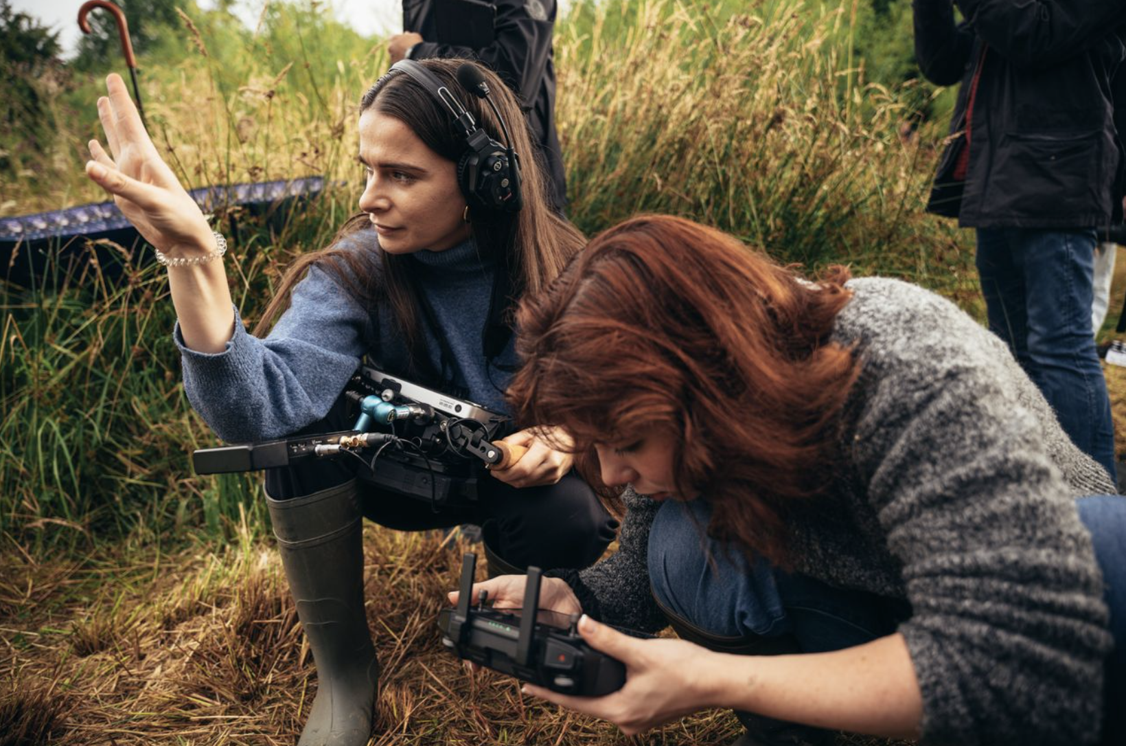 Two women working outdoors, one with a headset and a drone controller, pointing into the distance, and the other focusing on a drone controller, surrounded by tall grass and a third person in the background.