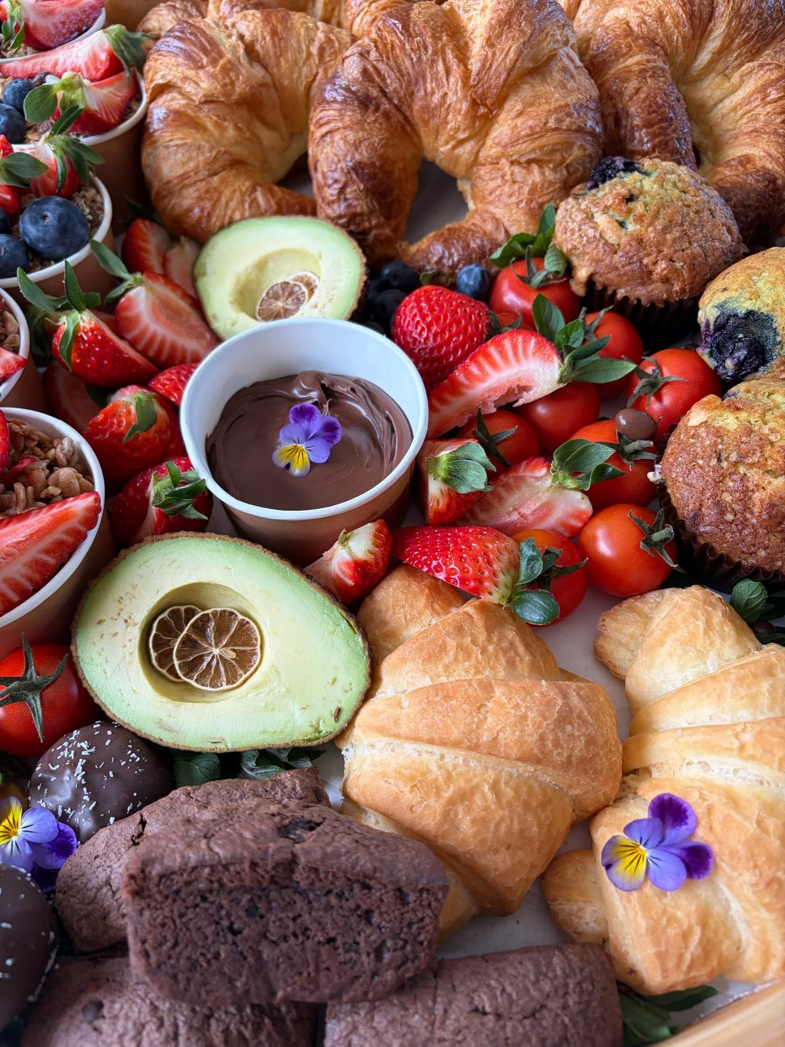 An assortment of breakfast items including croissants, muffins, strawberries, tomatoes, avocado with lemon slices, chocolate and vanilla pastries, and a cup of chocolate pudding garnished with edible flowers.