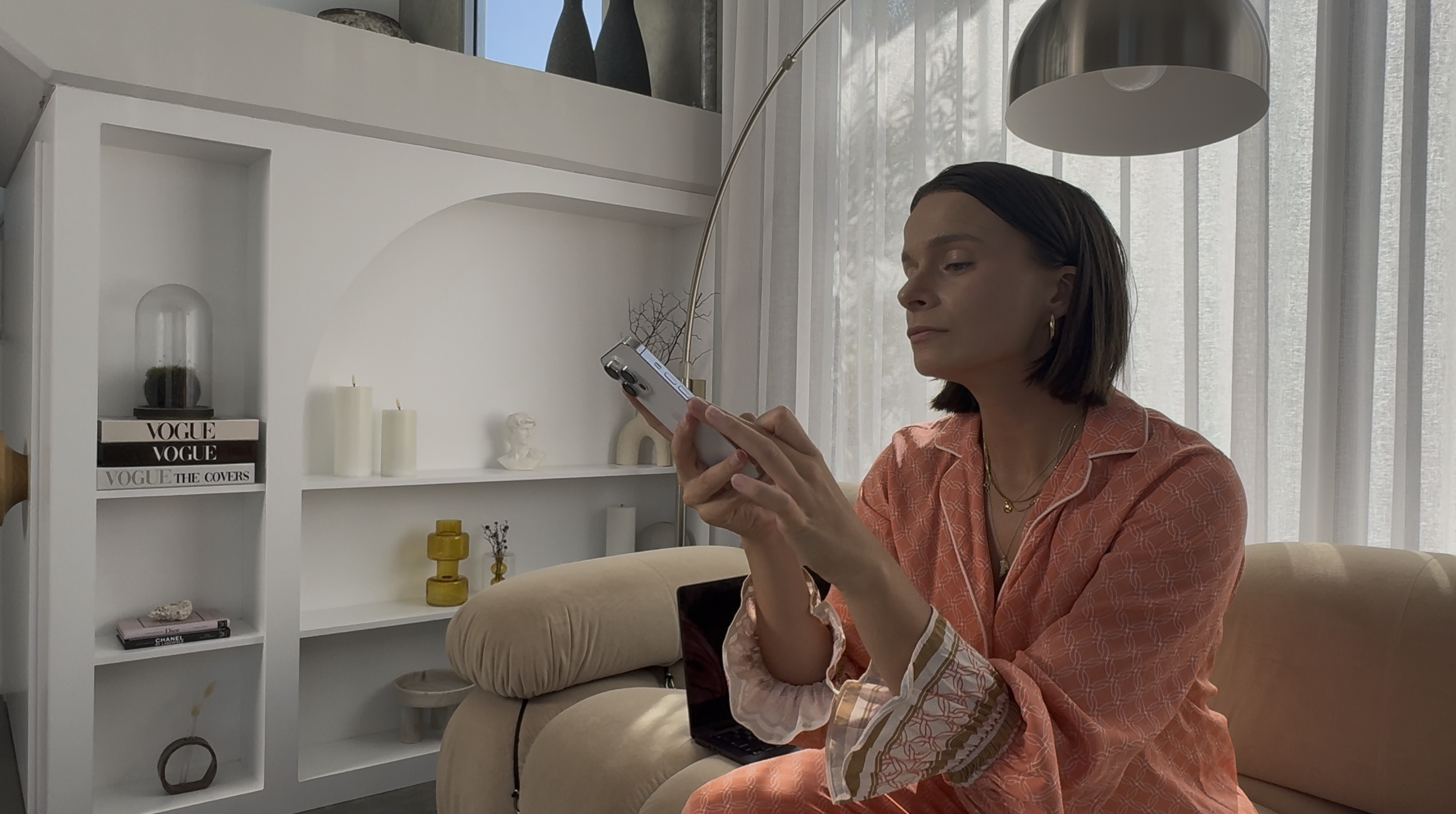 Woman sitting on a beige sofa, wearing an orange patterned shirt, using her smartphone, with a white bookshelf behind her displaying books and decorative items, and sheer curtains covering a window.