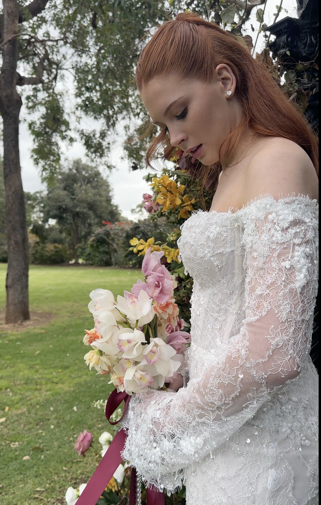 A bride in an off-shoulder lace wedding dress holding a bouquet of pink and white flowers outdoors.