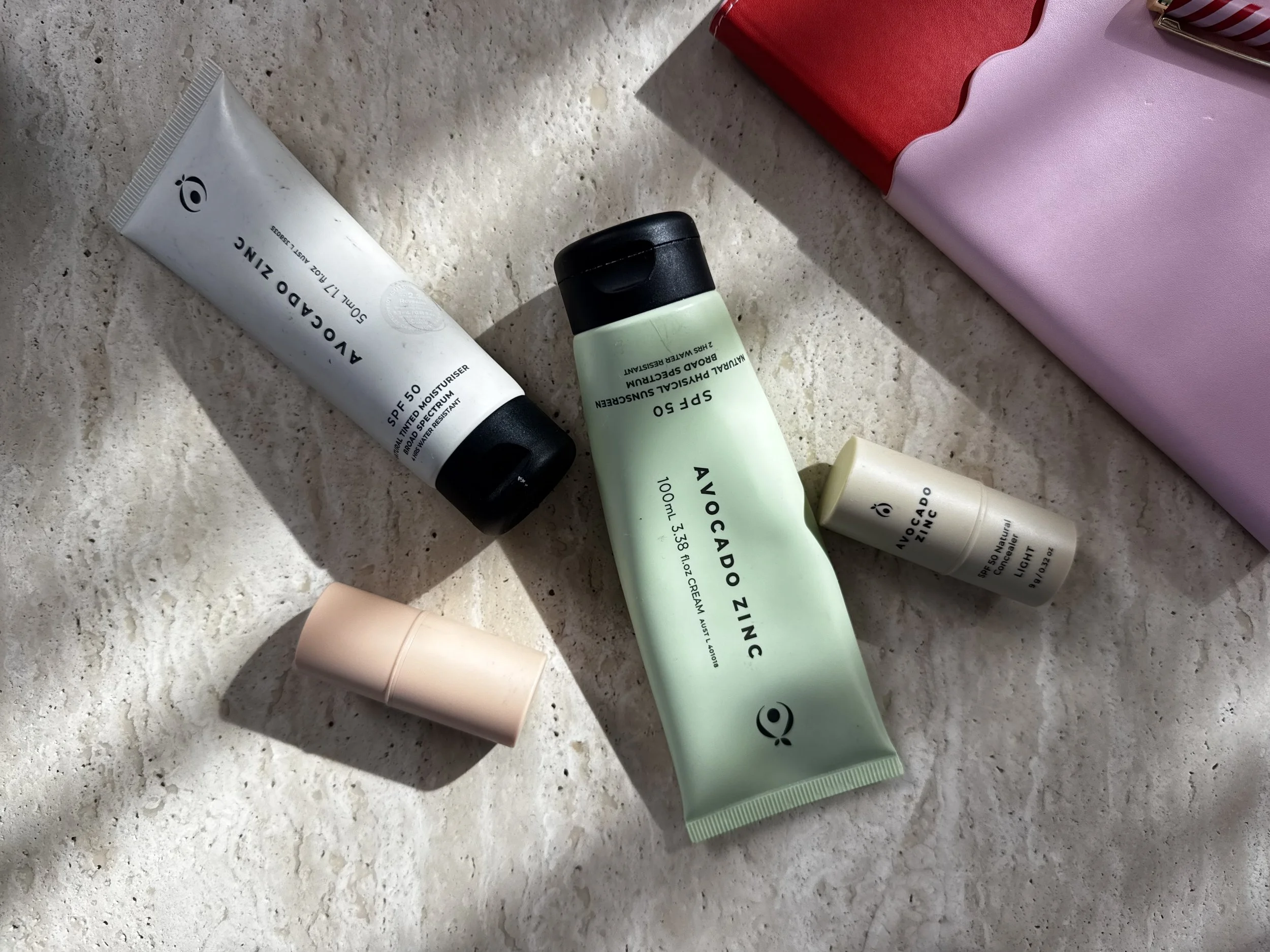 Assorted skincare and cosmetic products on a tiled surface, including a green tube labeled 'Avocado Zinc' sun cream, white tube labeled 'Autoado Zinc' sunscreen, small beige bottle, pink cylindrical container, and a pink bag in the upper right corner.