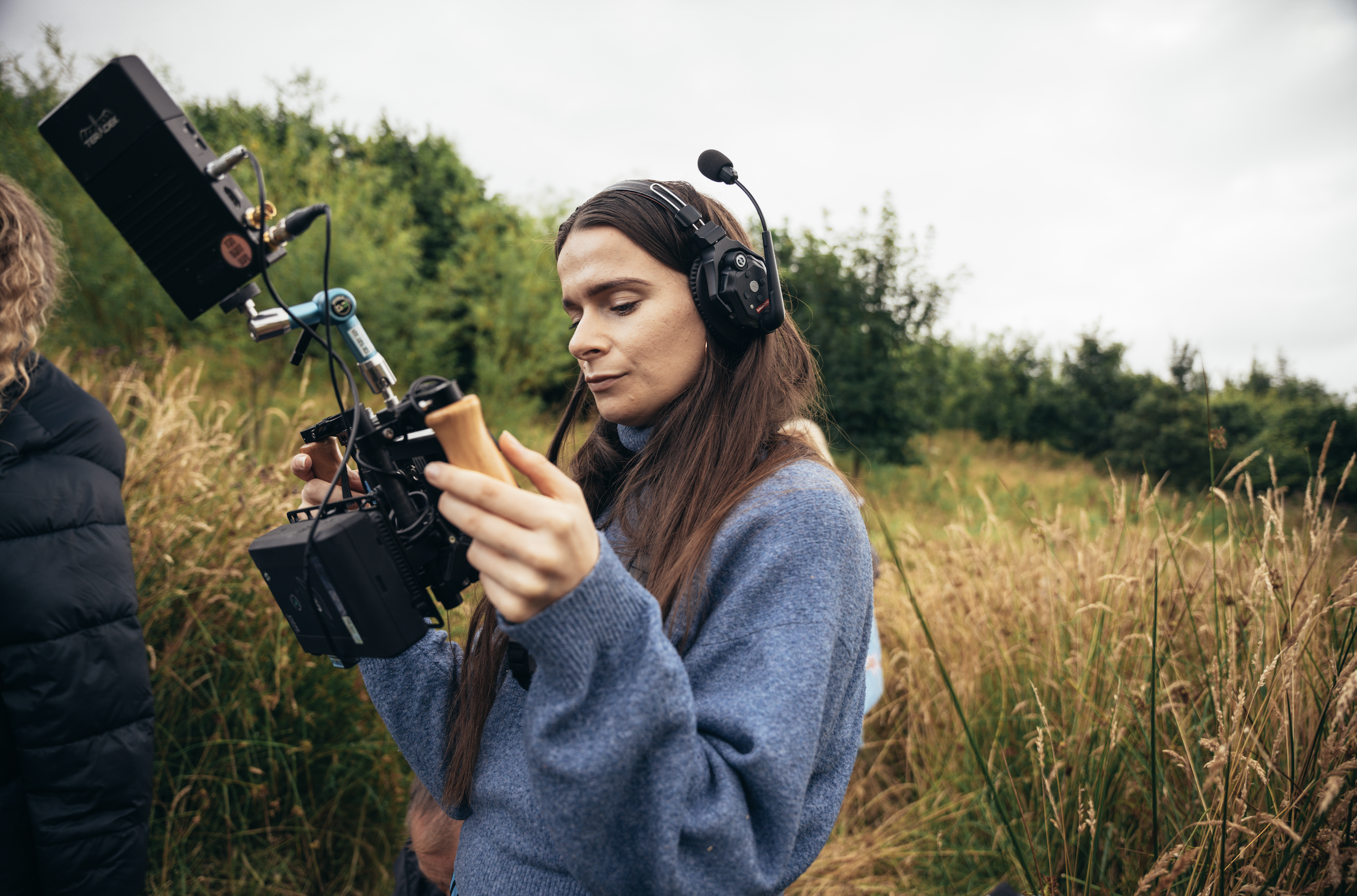 A woman with long brown hair, wearing a blue sweater and over-ear headphones, looking at a camera mounted on a stabilizer in an outdoor field with tall grass and trees in the background.