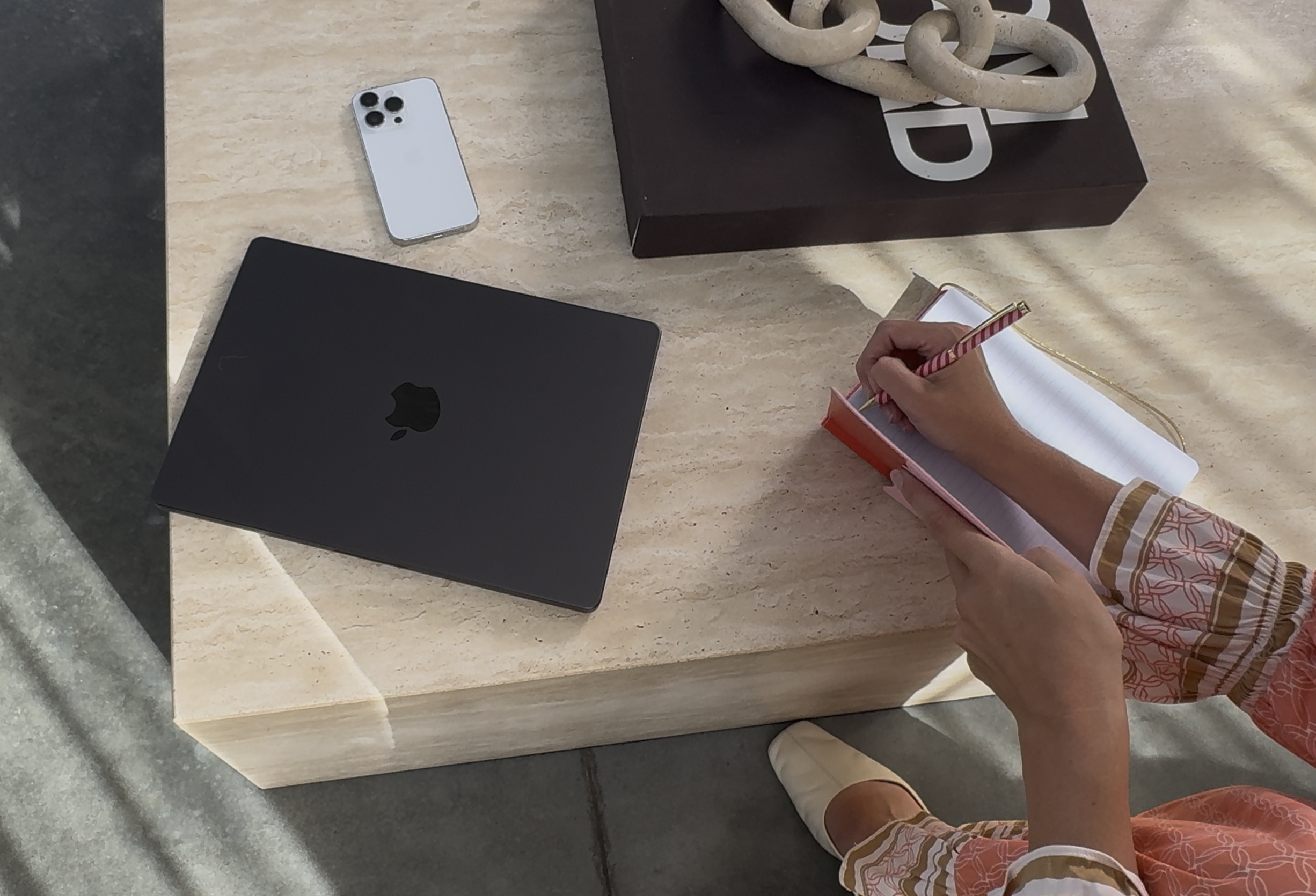 A person is sitting at a table writing in a notebook with a striped pen. On the table, there is a closed black Apple MacBook, a white iPhone, and a large brown box with decorative loops on top. The person's outfit has a colorful, patterned fabric.