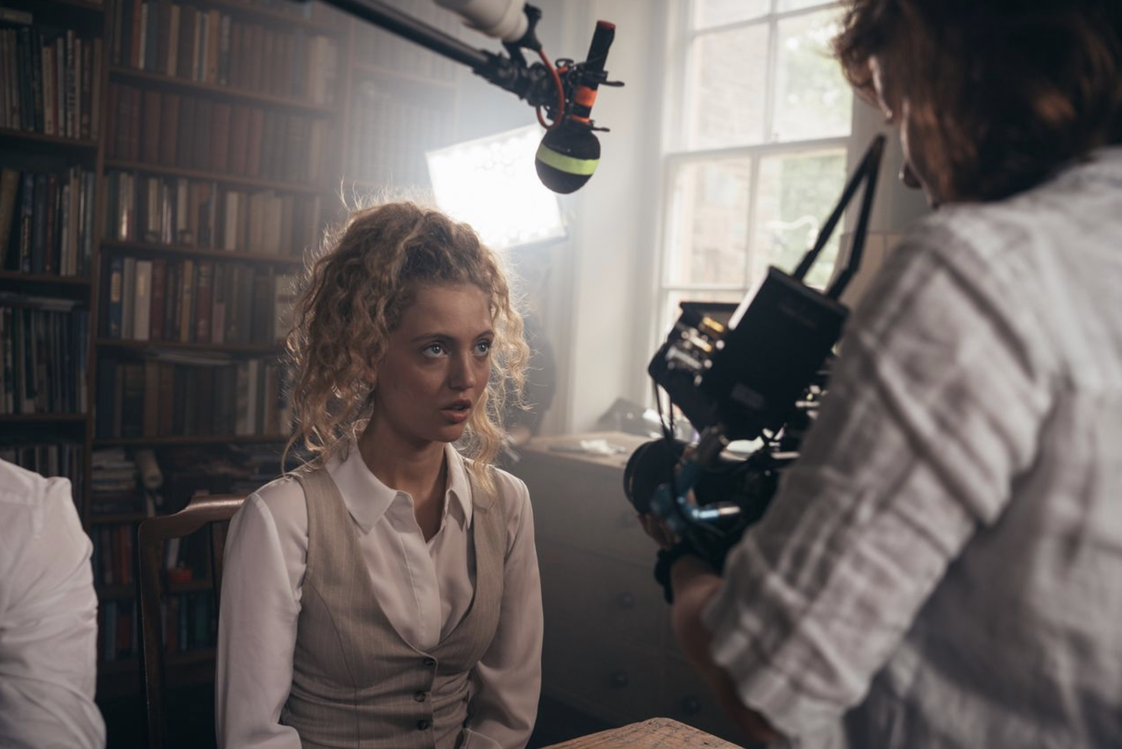 A woman with curly blonde hair being filmed by a crew member holding a camera in a room with a bookshelf and window, during daylight.