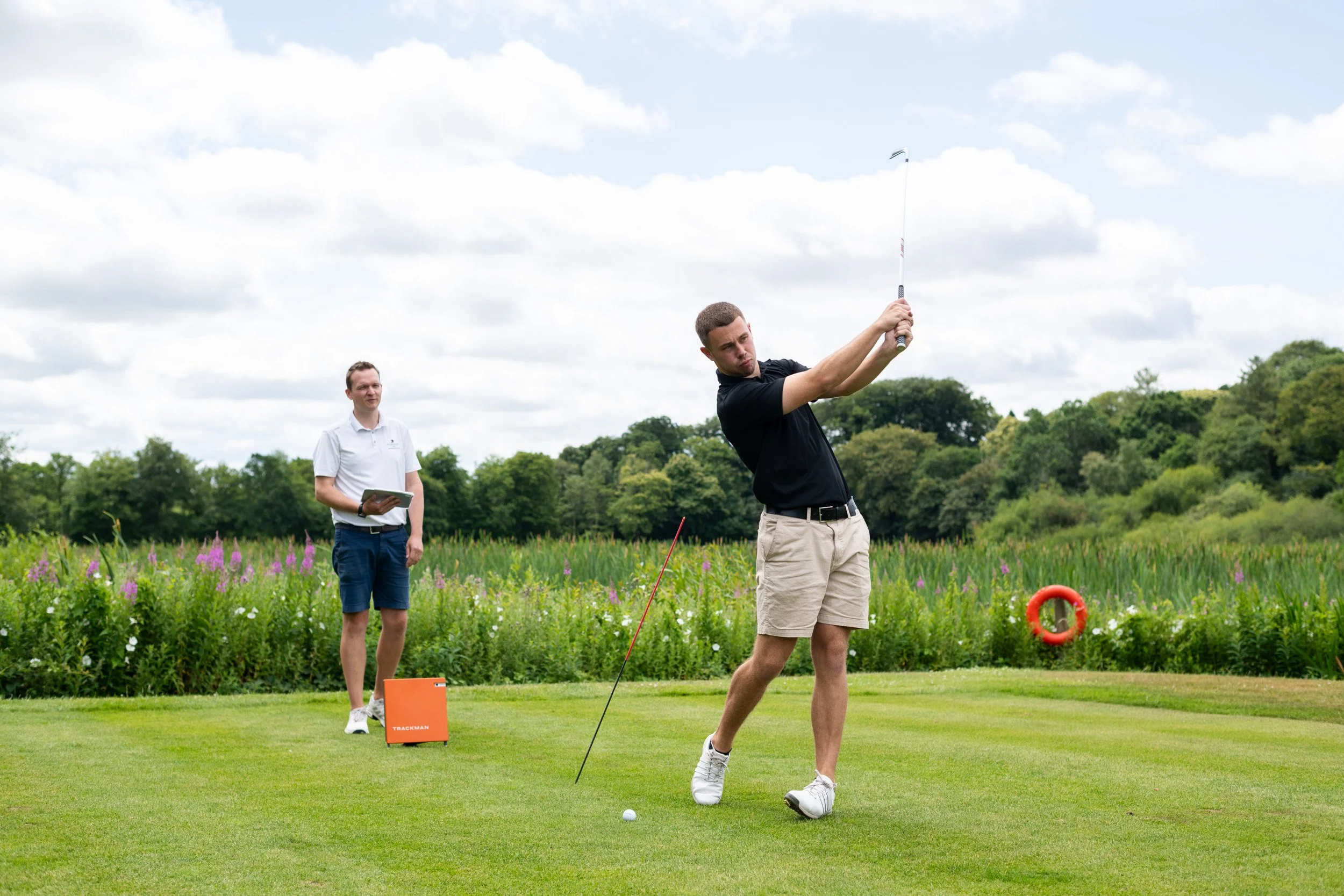 A man in a black shirt and beige shorts swings a golf club on a golf course while another man in a white shirt and blue shorts observes, holding a clipboard. There is a golf ball on the grass and a red lifebuoy on a pole in the background, with lush green trees and a partly cloudy sky.