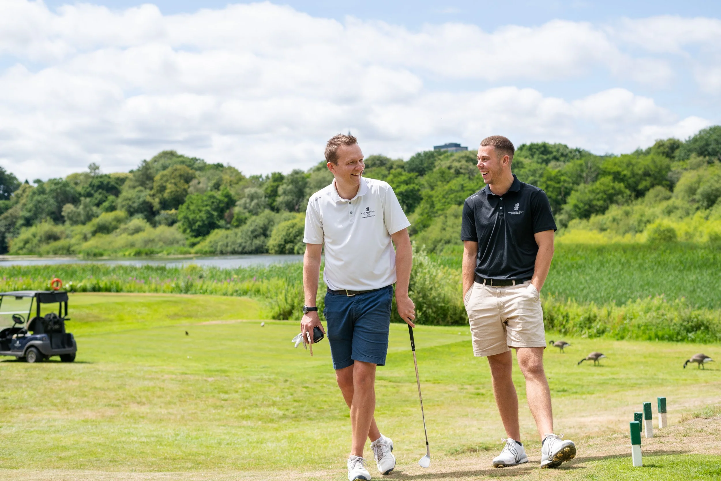 Two men walking and talking on a golf course, dressed in golf attire, with a golf cart in the background, lush greenery, and a partly cloudy sky.