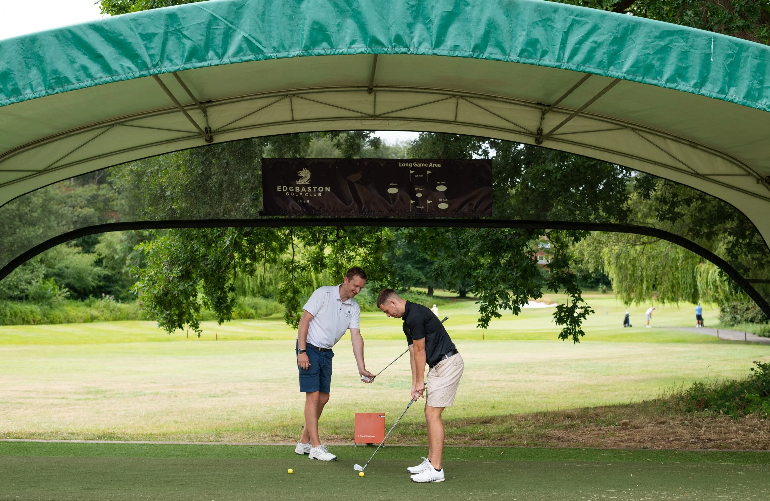 Two men at a golf driving range, one with a black shirt and beige shorts, the other with a white shirt and blue shorts, practicing their golf swings under a large green canopy with a sign for Edgbaston Golf Club. In the background, there is lush green grass and several people playing golf.