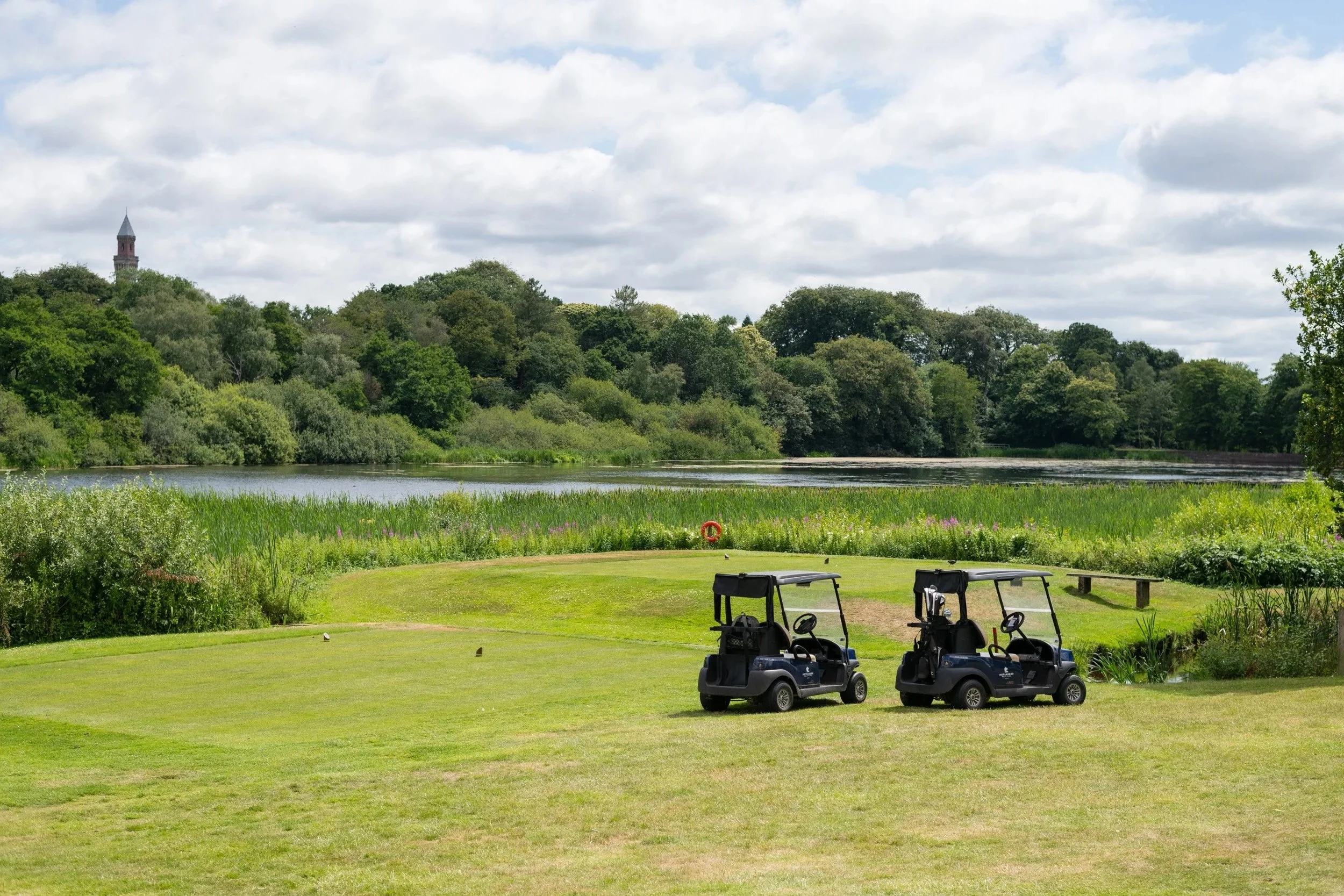 Two golf carts parked on a grassy area near a pond with lush trees in the background, under a partly cloudy sky.