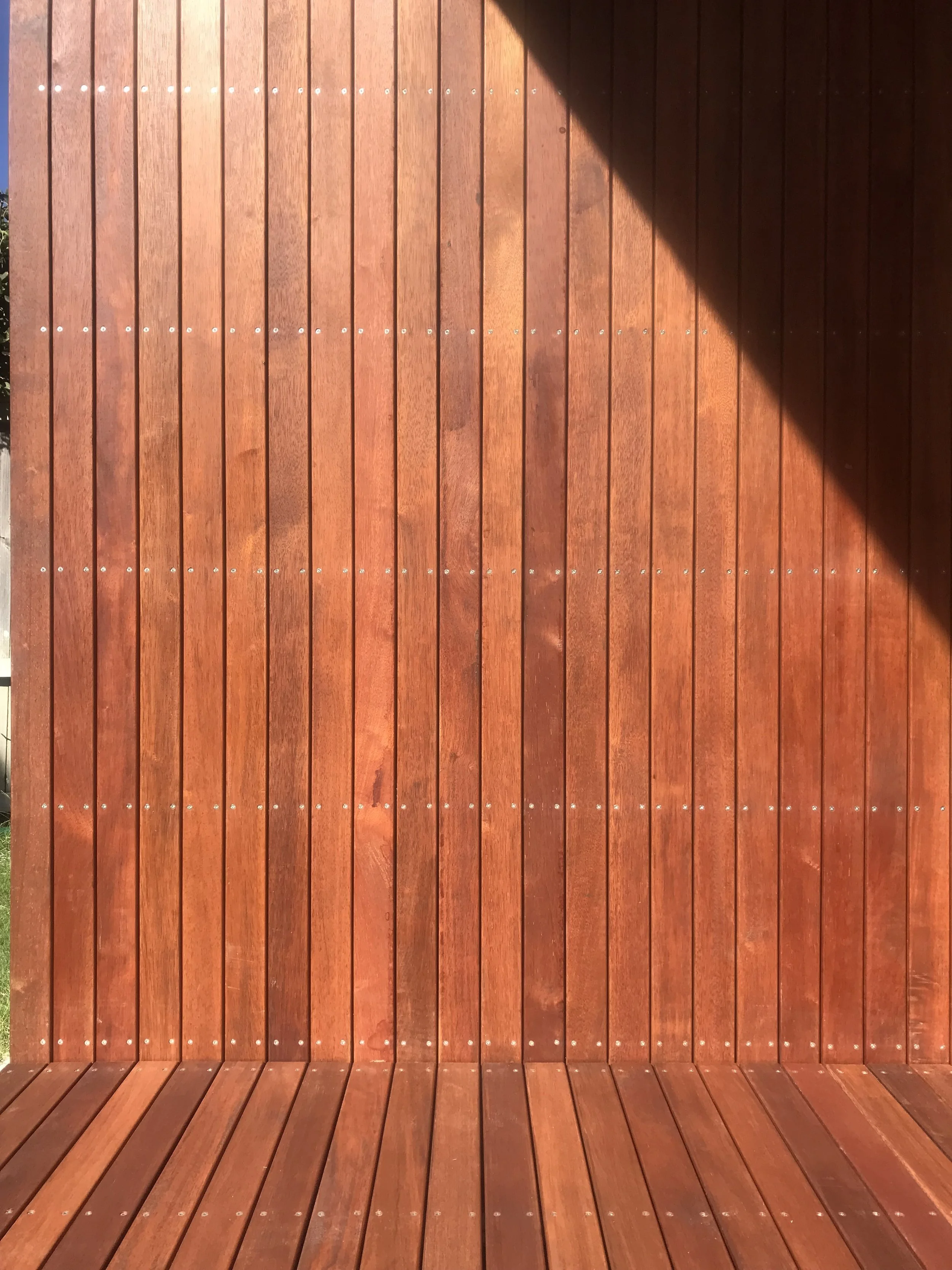 Wooden outdoor structure with vertical and horizontal planks, sunlit with a shadow across the top right corner.