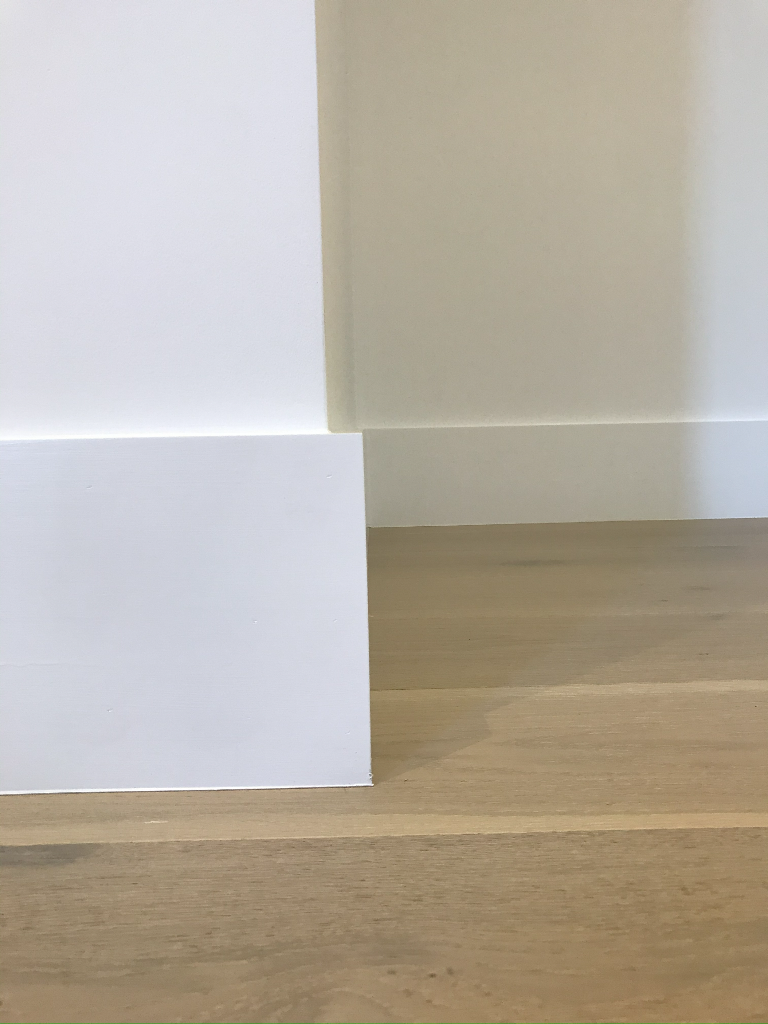 Close-up of the corner of a wooden floor, with white and beige walls meeting at a right angle, and a white baseboard along the wall.
