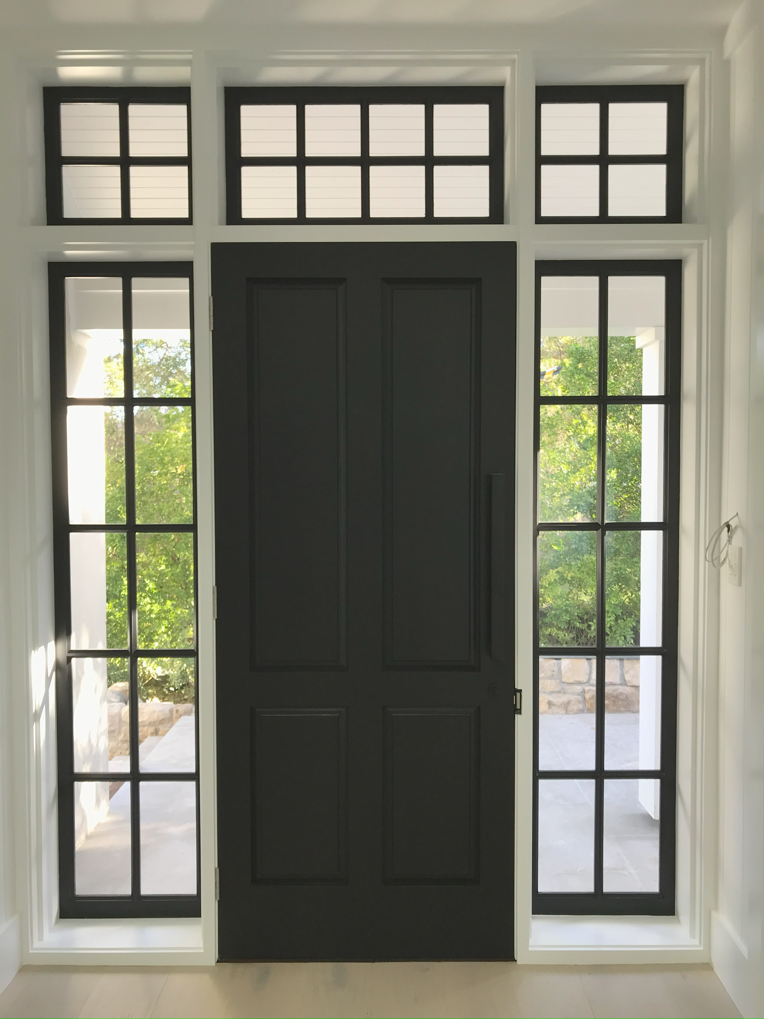 Interior view of a front door with black framing, surrounded by large glass windows and transom windows above, showing a porch and greenery outside.