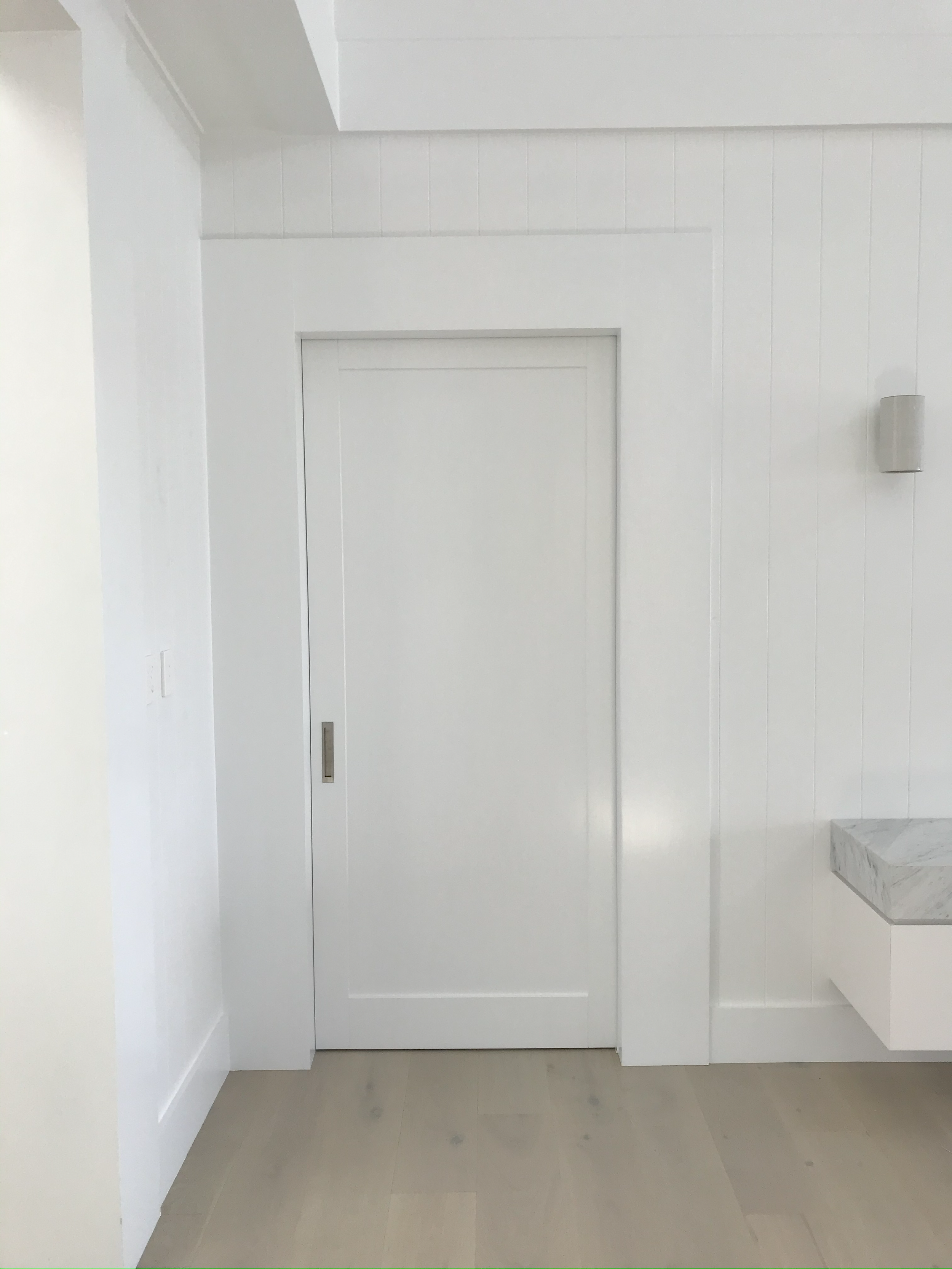 Interior view of a white door with a handle, surrounded by white wall paneling and baseboards, with a light-colored wooden floor and part of a marble countertop visible on the right.