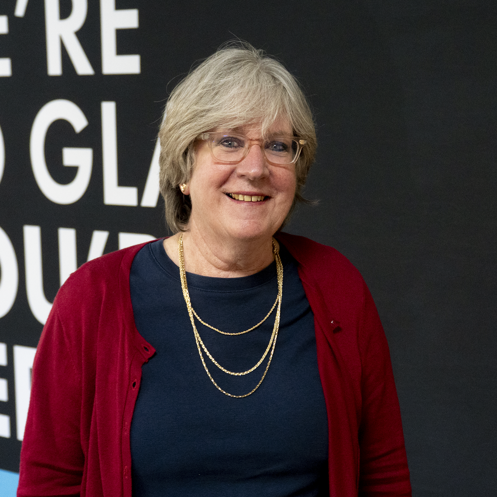 A smiling woman with short gray hair, glasses, and gold jewelry, wearing a dark top and a red cardigan, standing in front of a black background with white text.