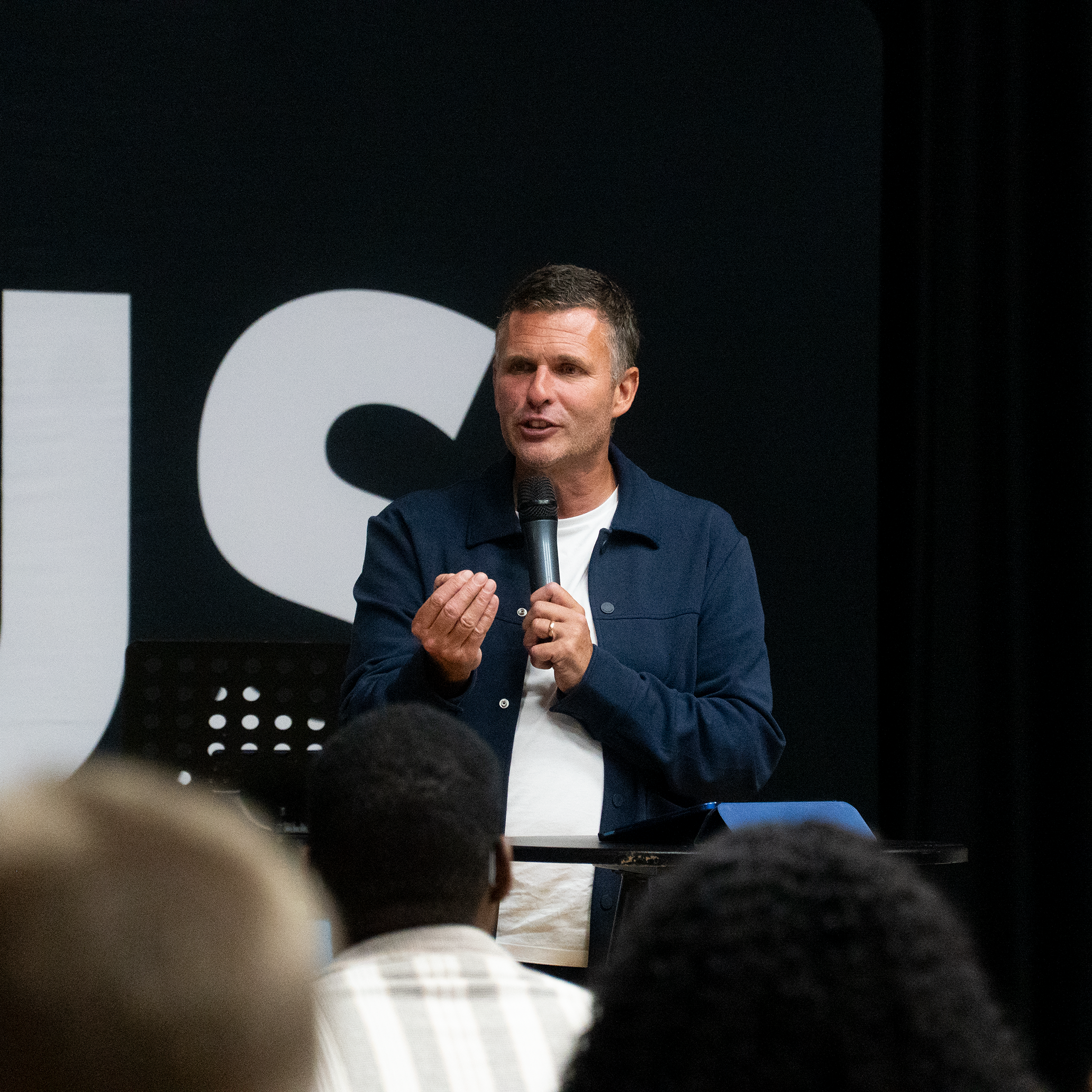 A man holding a microphone and speaking at a presentation or event, with a large black and white background in the style of a speaker or conference setting.
