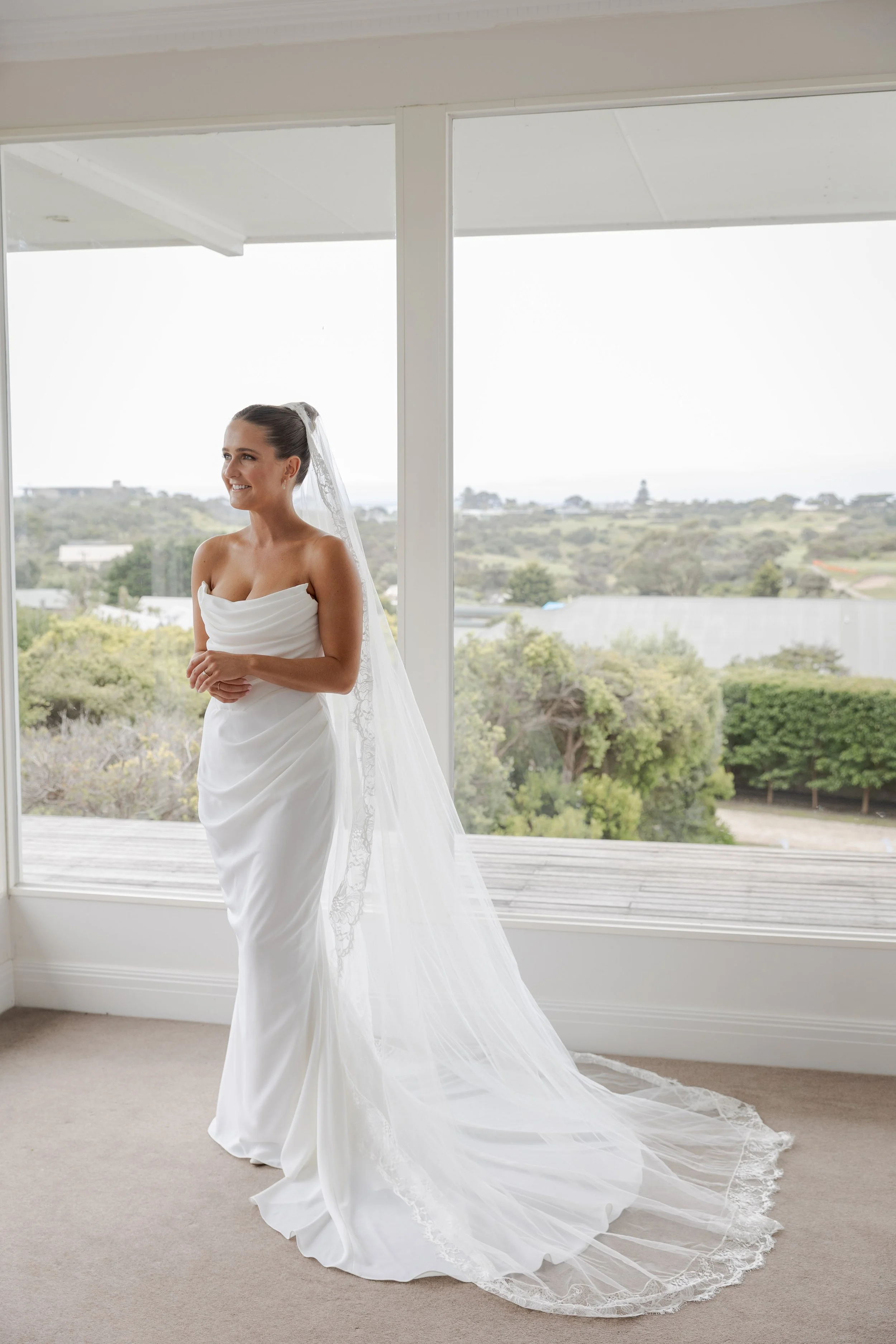 A bride smiles as she stands in her bedroom, dressed in her wedding dress and veil, moments before she walks down the aisle in Portsea, Melbourne.