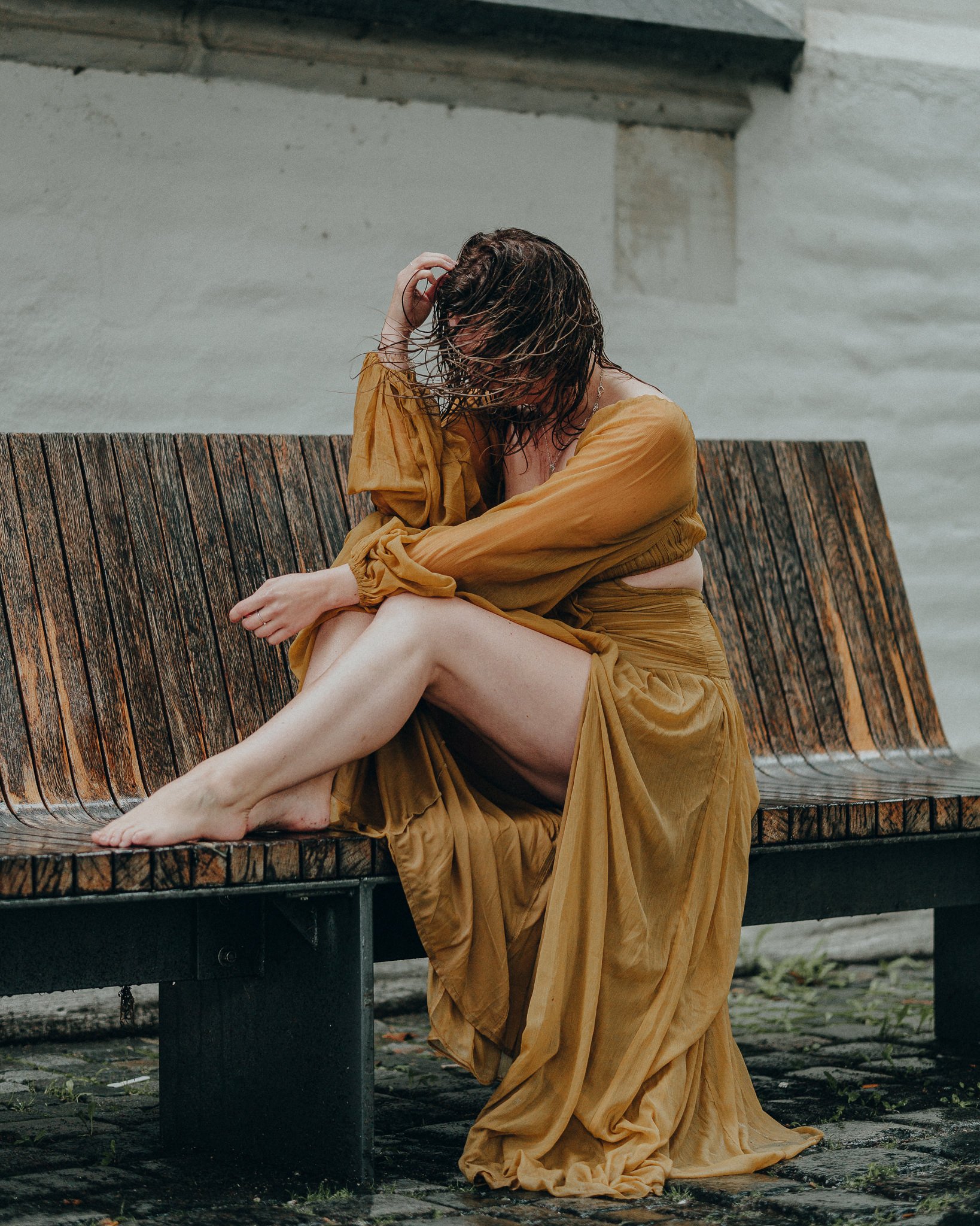 Eine Frau in einem langen, goldgelben Kleid sitzt auf einer Holzbank, die nasse Haare hat und scheinen vom Regen nass zu sein.