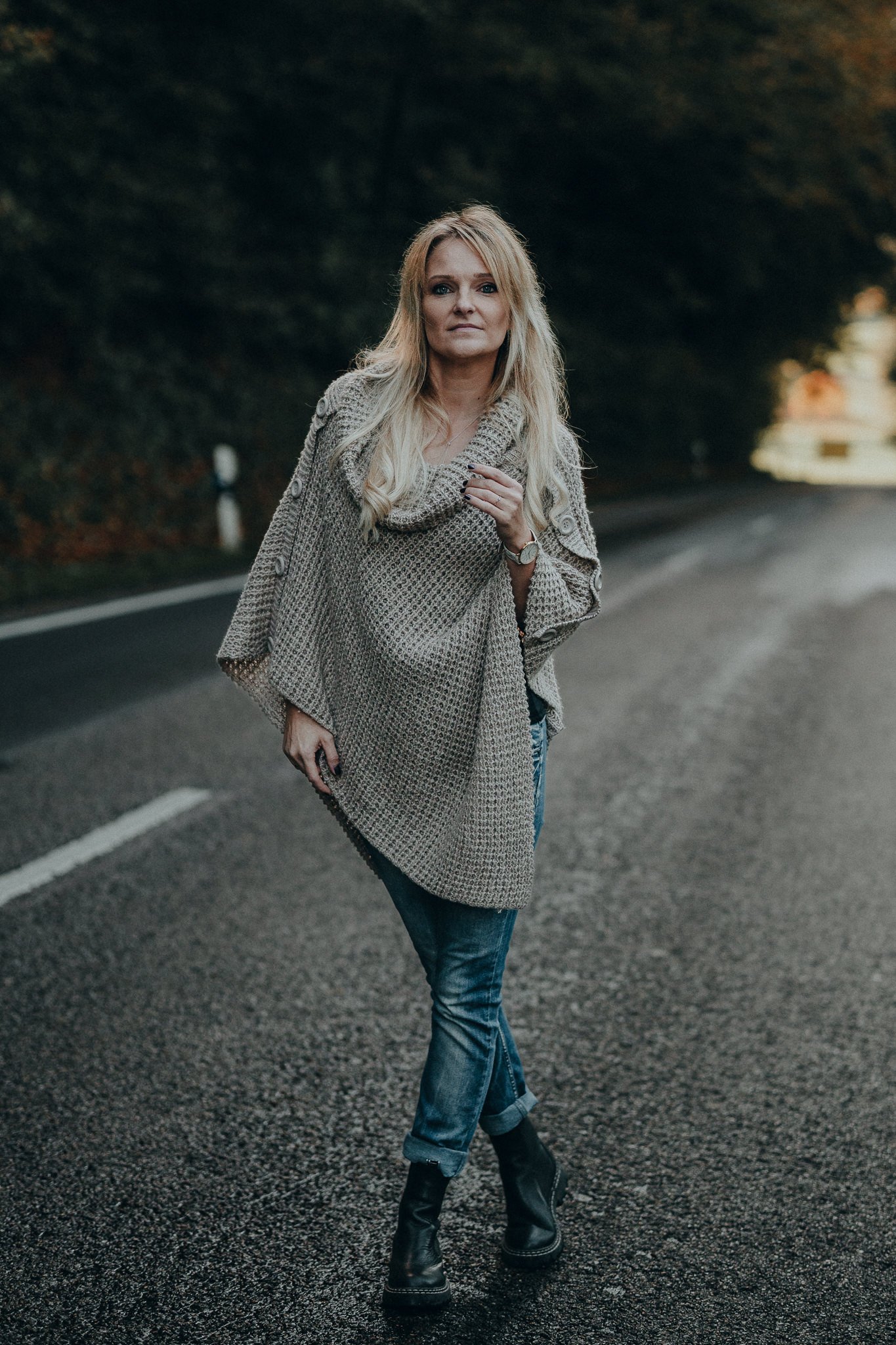 Eine blonde Frau in einer großen, grauen Strickjacke, Jeans und schwarzen Stiefeln steht auf einer dunklen, leeren Strasse bei Dämmerung im Freien.