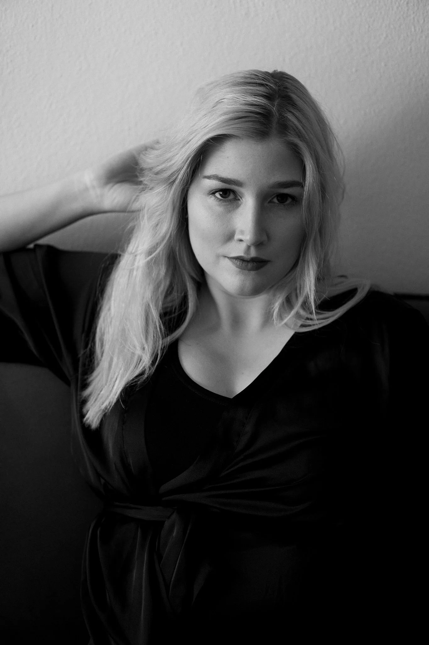 Schwarz-weiß-Fotografie einer jungen Frau mit langem, leicht gewelltem Haar, sitzend gegen die Wand, mit nachdenklichem Blick und legerer Haltung.