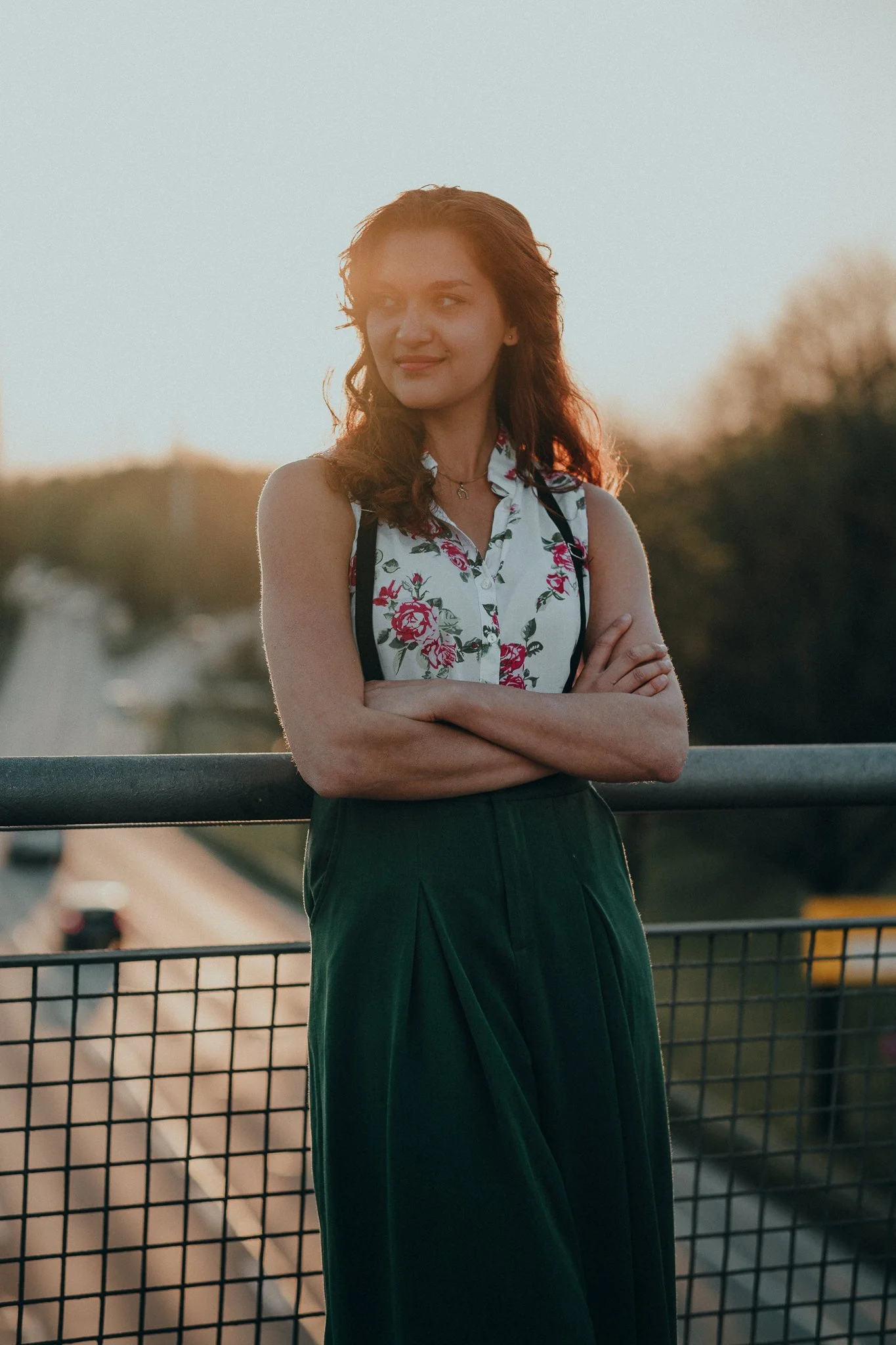Eine junge Frau steht auf einer Brücke bei Sonnenuntergang, trägt ein ärmelloses Blusen-Shirt mit Rosenmuster und ein dunklen Rock, mit verschränkten Armen und Blick in die Ferne.
