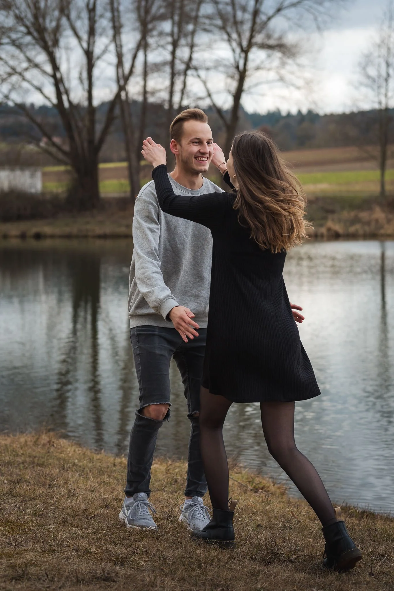 Ein lachendes Paar tanzt am Ufer eines Sees im Freien, umgeben von Bäumen und einer ländlichen Landschaft.