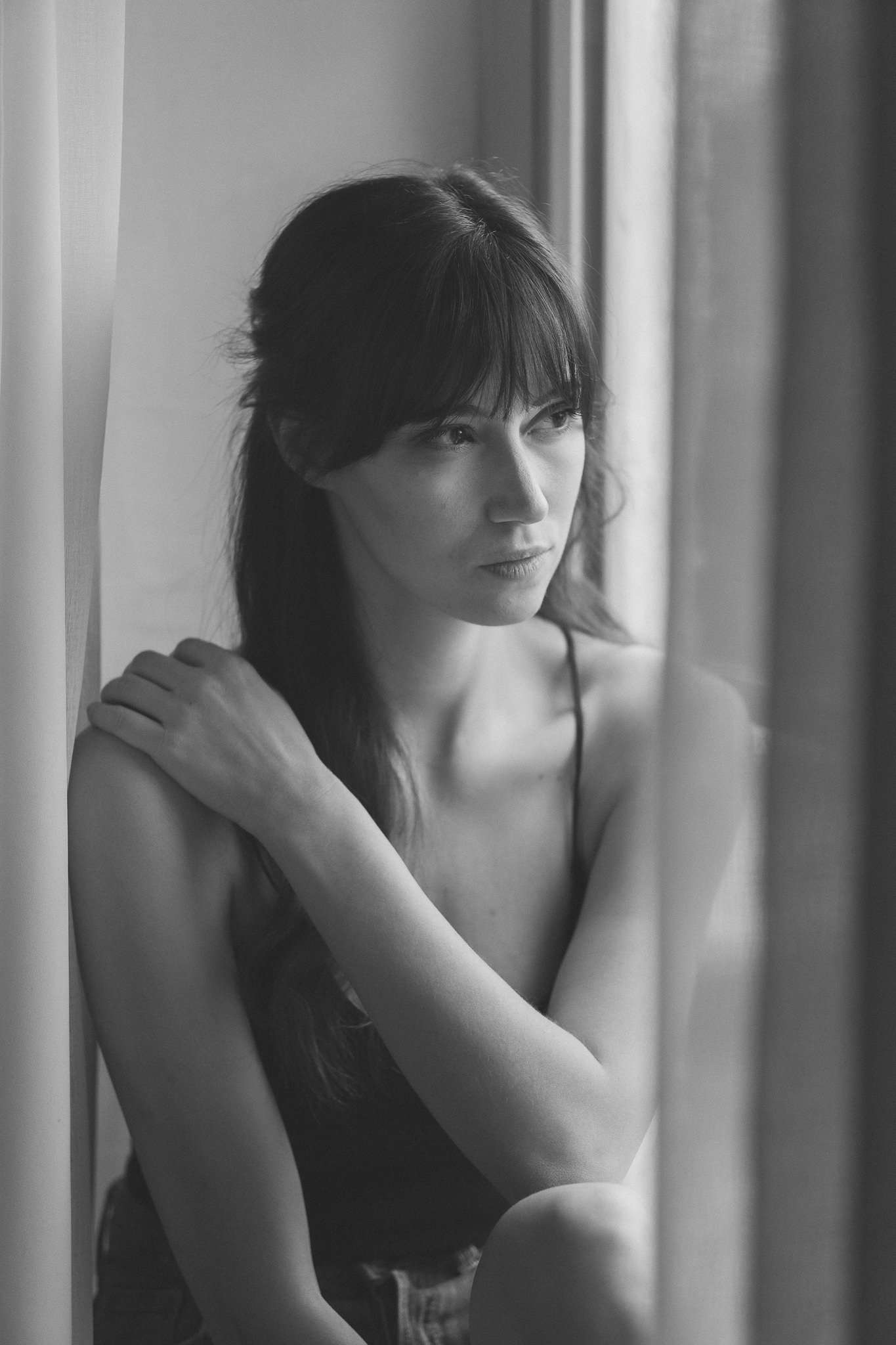 Schwarz-weiß-Fotografie einer jungen Frau mit dunkel aussehendem Haar, die nach draußen schaut. Sie trägt ein dünnes, schwarzes Top und sitzt neben einem Fenster, das sanftes Licht auf ihr Gesicht wirft.