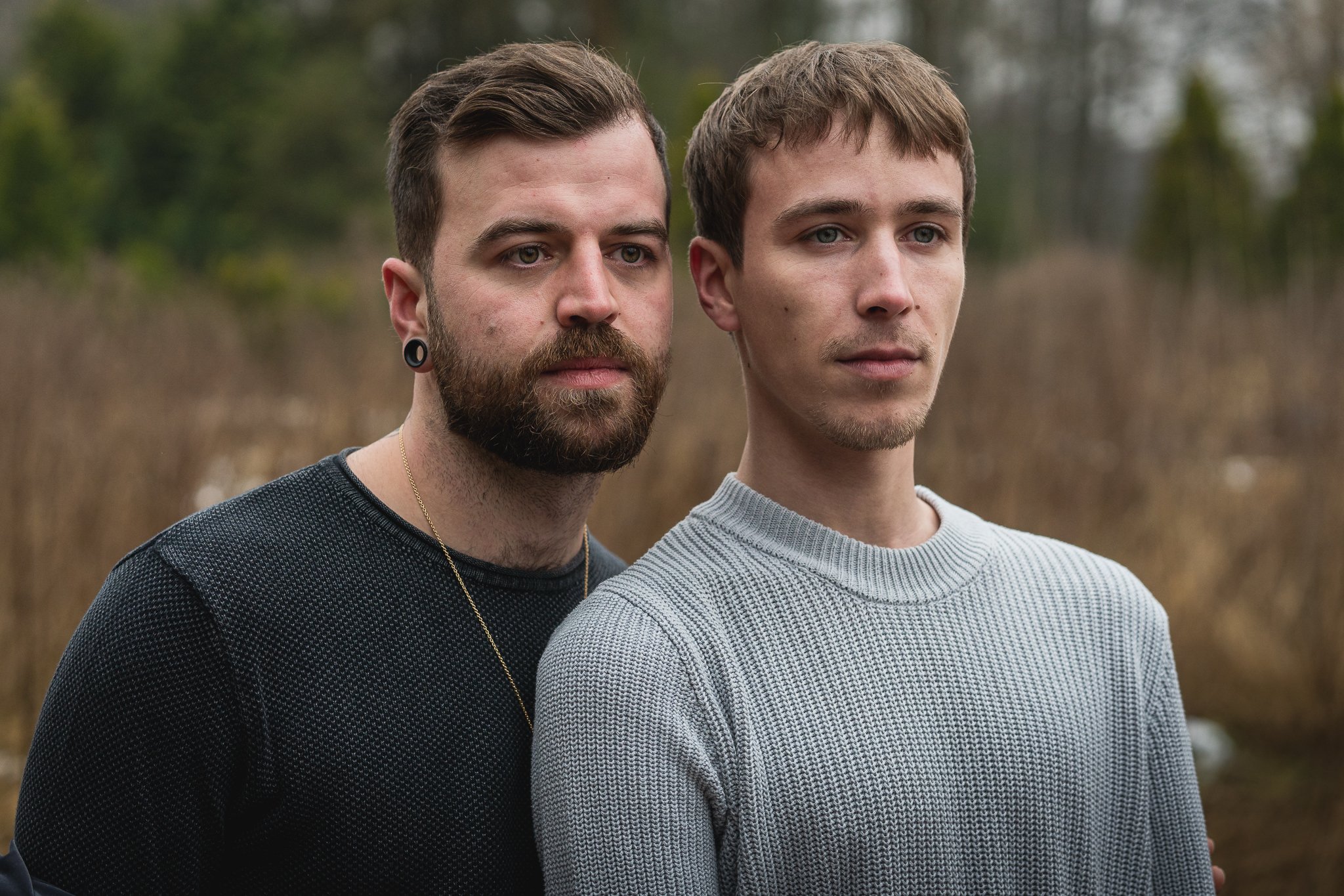 Zwei junge Männer stehen draußen in einer natürlichen Umgebung, umgeben von Bäumen und einem braunen Grasland, mit ernsten Ausdrücken auf ihren Gesichtern.
