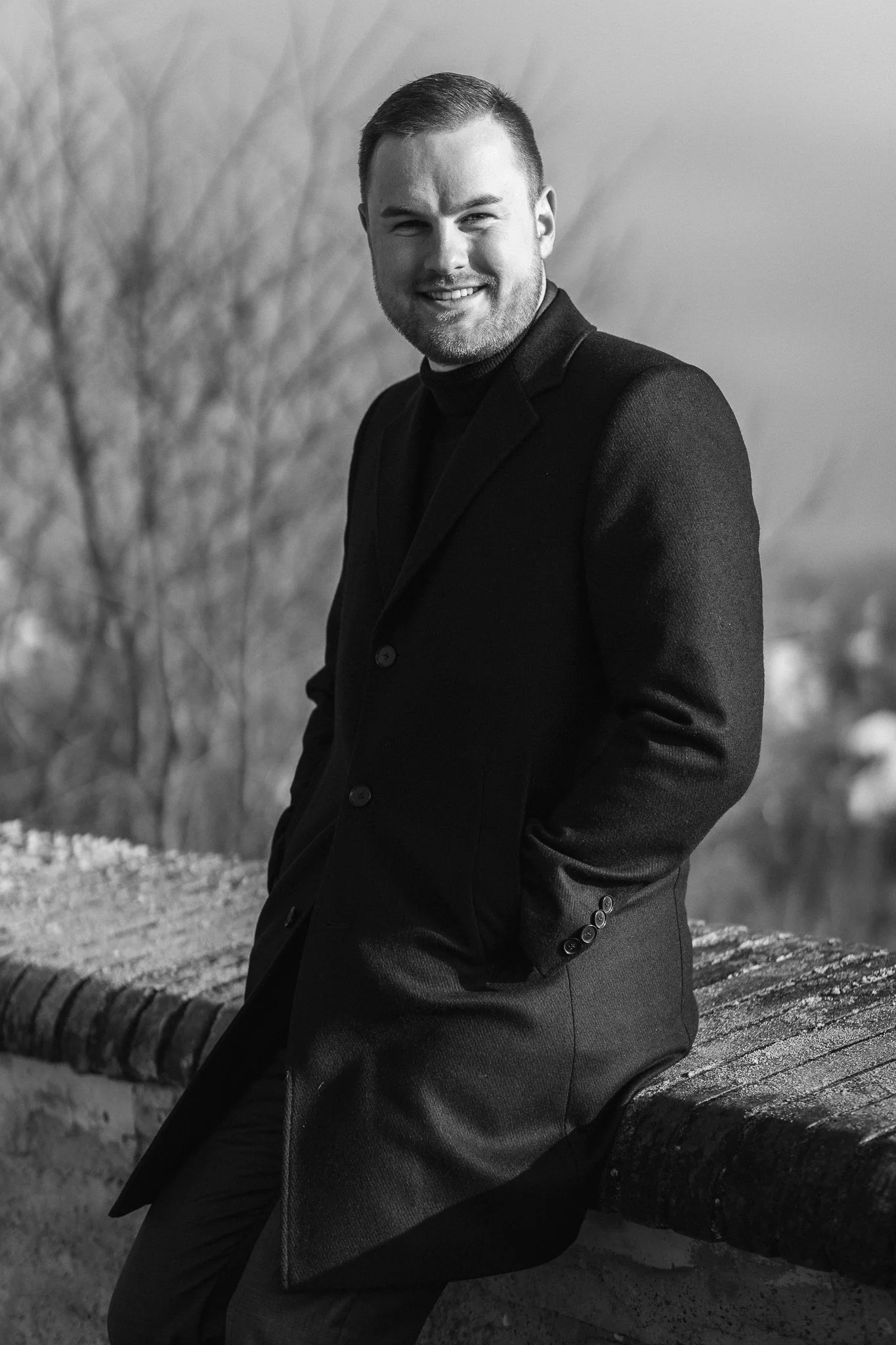 Schwarz-Weiß-Fotografie eines lächelnden Mannes in einem dunklen Anzug, der an einer Mauer im Freien lehnt.