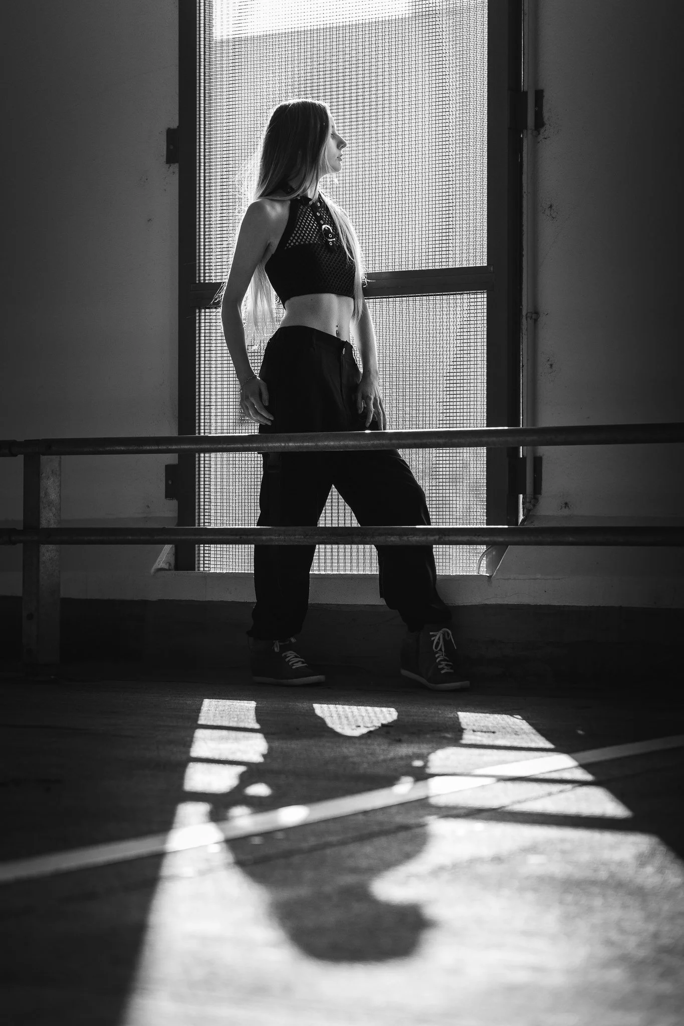 Eine junge Frau steht an einem Fenster, fotografiert in Schwarz-Weiß, mit Schatten auf dem Boden, trägt Sportkleidung und Sneaker.