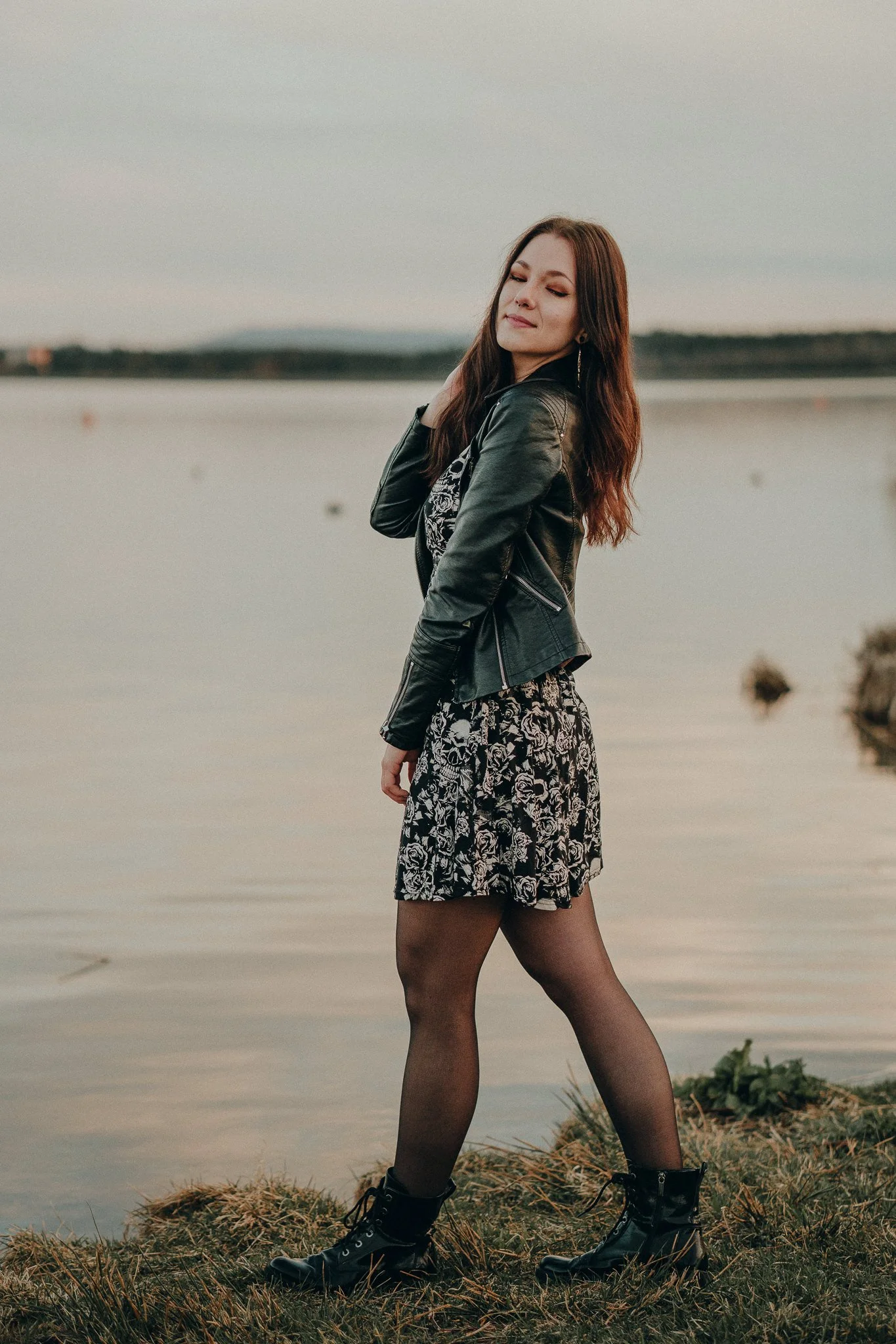 Junge Frau am Wasser bei Sonnenuntergang, trägt Gummistiefel, schwarzes Kleid mit Blümchenmuster und Lederjacke.