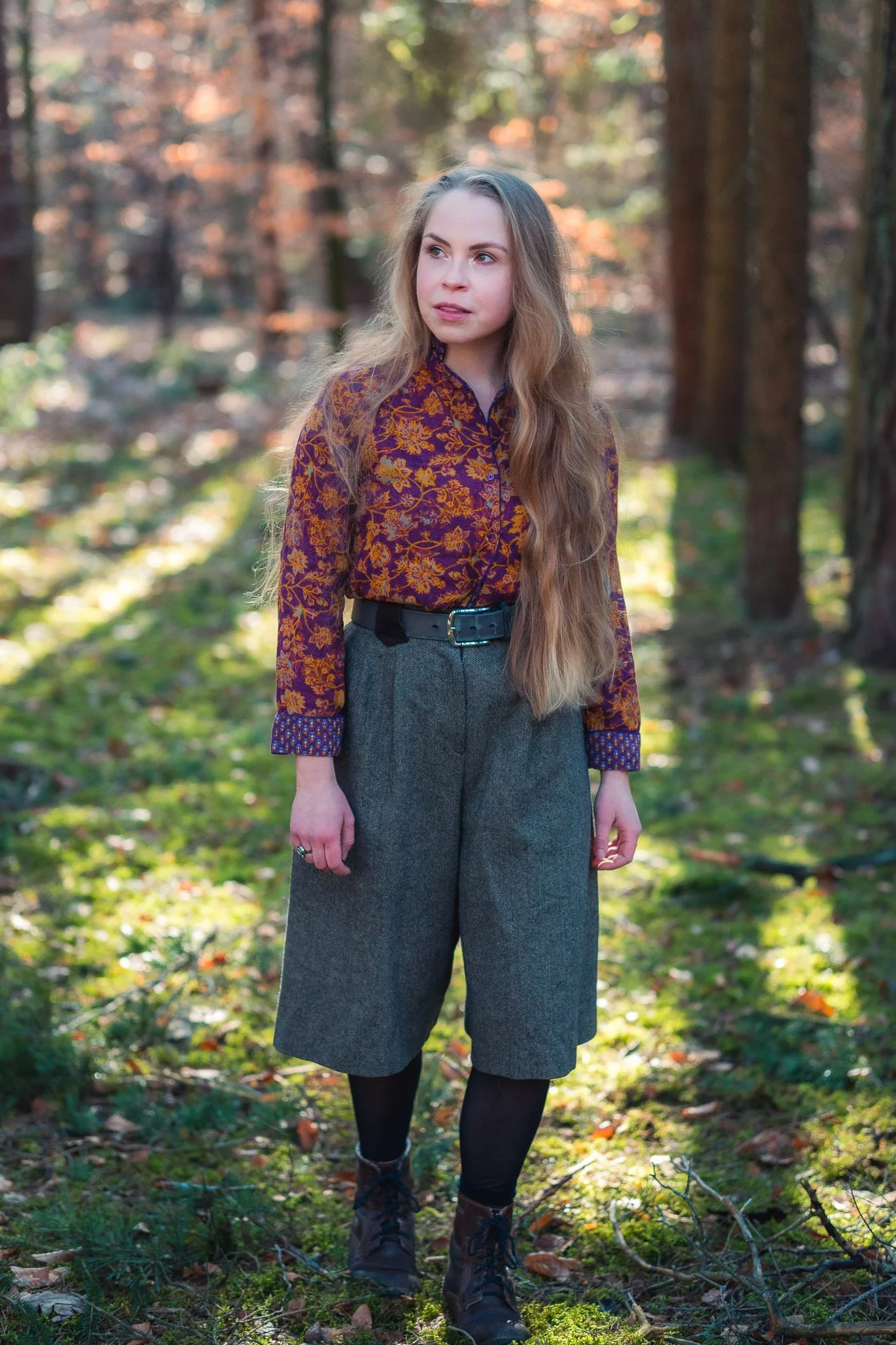 Eine junge Frau mit langen, gewellten braunen Haaren steht in einem Wald im Herbst. Sie trägt eine bunte, florale Bluse, graue Hosen und robuste Stiefel.