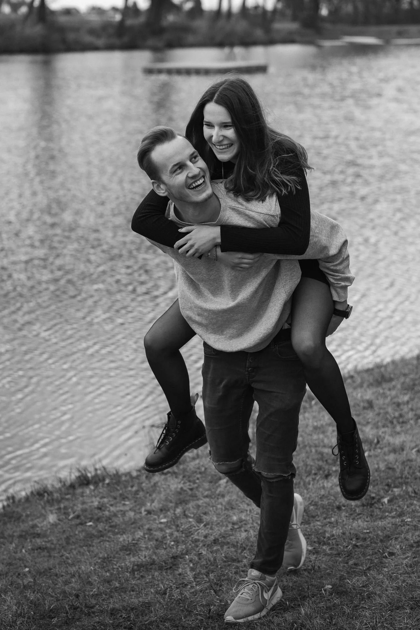 Ein junges Paar spielt lachend am Flussufer, der Mann trägt ein Sweatshirt, das Mädchen sitzt auf seinen Schultern, beide wirken fröhlich.