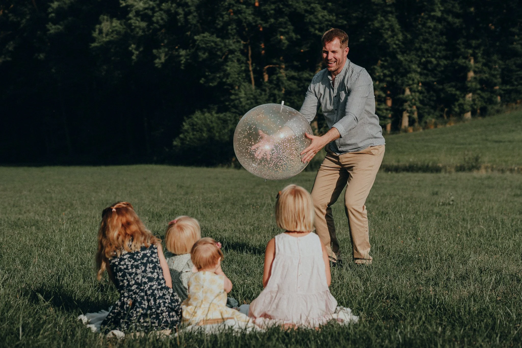 Ein Erwachsener spielt mit einem großen transparenten Ball mit Glitzer und leuchtenden Lichtern, während vier Kinder auf einer Decke im Gras sitzen, in einer grünen, bewaldeten Landschaft bei Sonnenuntergang.