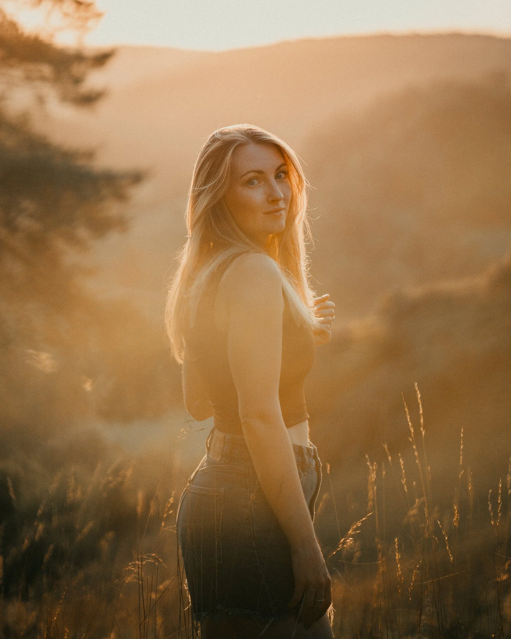 Eine junge Frau mit langem blondem Haar steht in einem Sonnenuntergang auf einer Wiese, umgeben von hohen Gräsern, im Hintergrund Hügel.