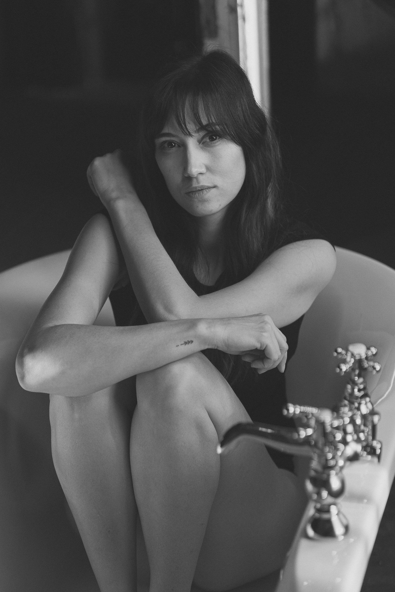 Schwarz-Weiß-Foto einer jungen Frau mit dunklen Haaren, die in einer Badewanne sitzt, die Beine angezogen, und die direkt in die Kamera schaut.