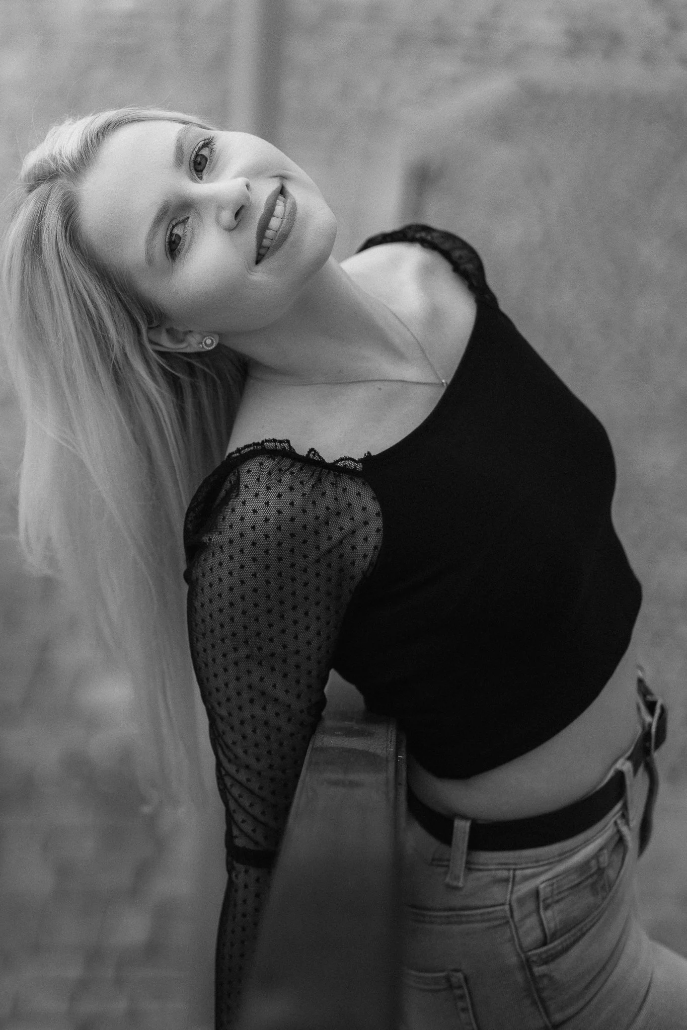 Eine lächelnde Frau mit langen blonden Haaren lehnt sich an eine Bar oder Theke. Sie trägt ein schwarzes Top mit durchsichtigen, gepunkteten Ärmeln und Jeans.