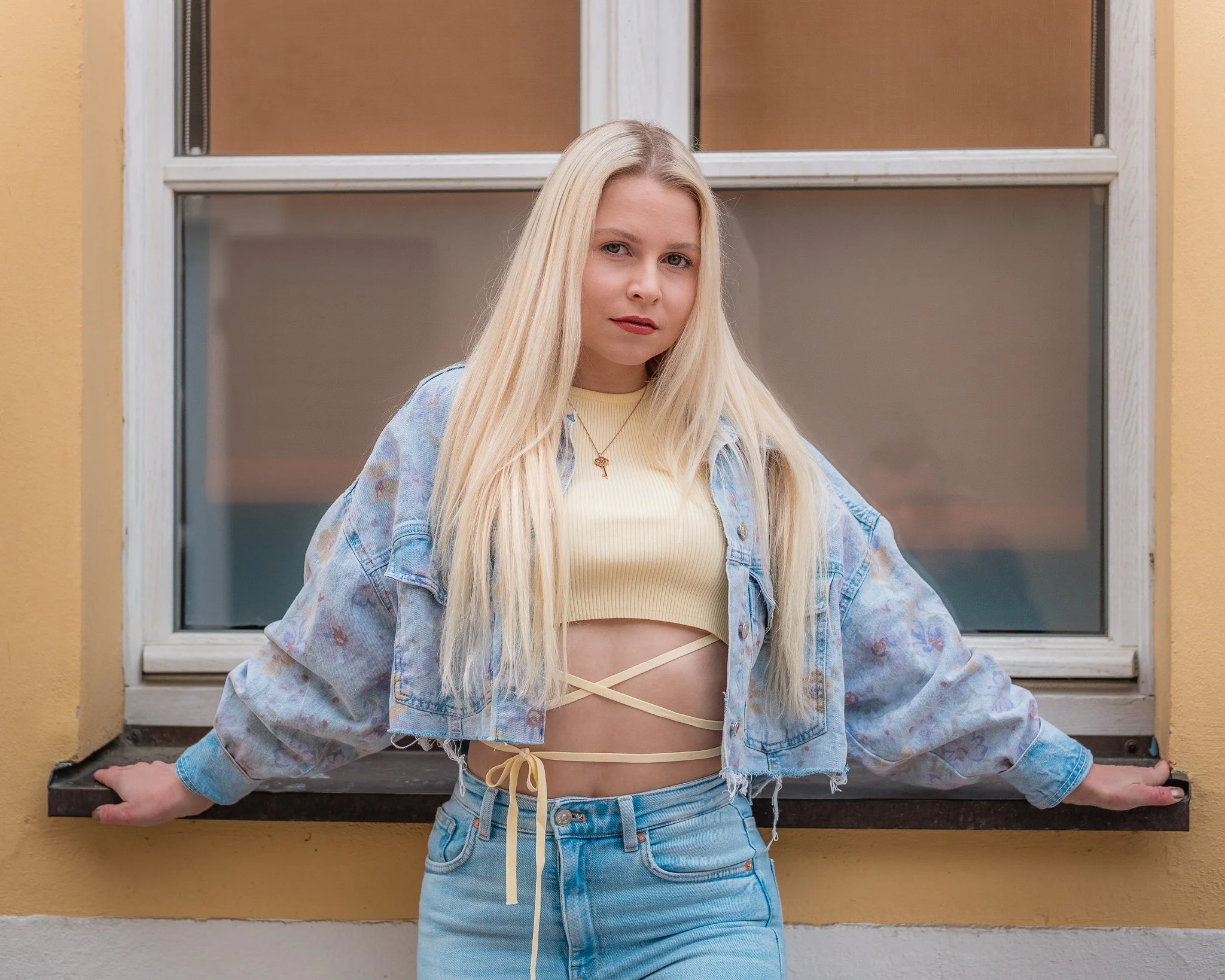 Junge Frau mit lange blondem Haar trägt eine gelbe Croptop, eine aufgeschnittene Lederjacke und Jeans, steht vor einem Fenster mit gelber Wand und Schwarztafel.