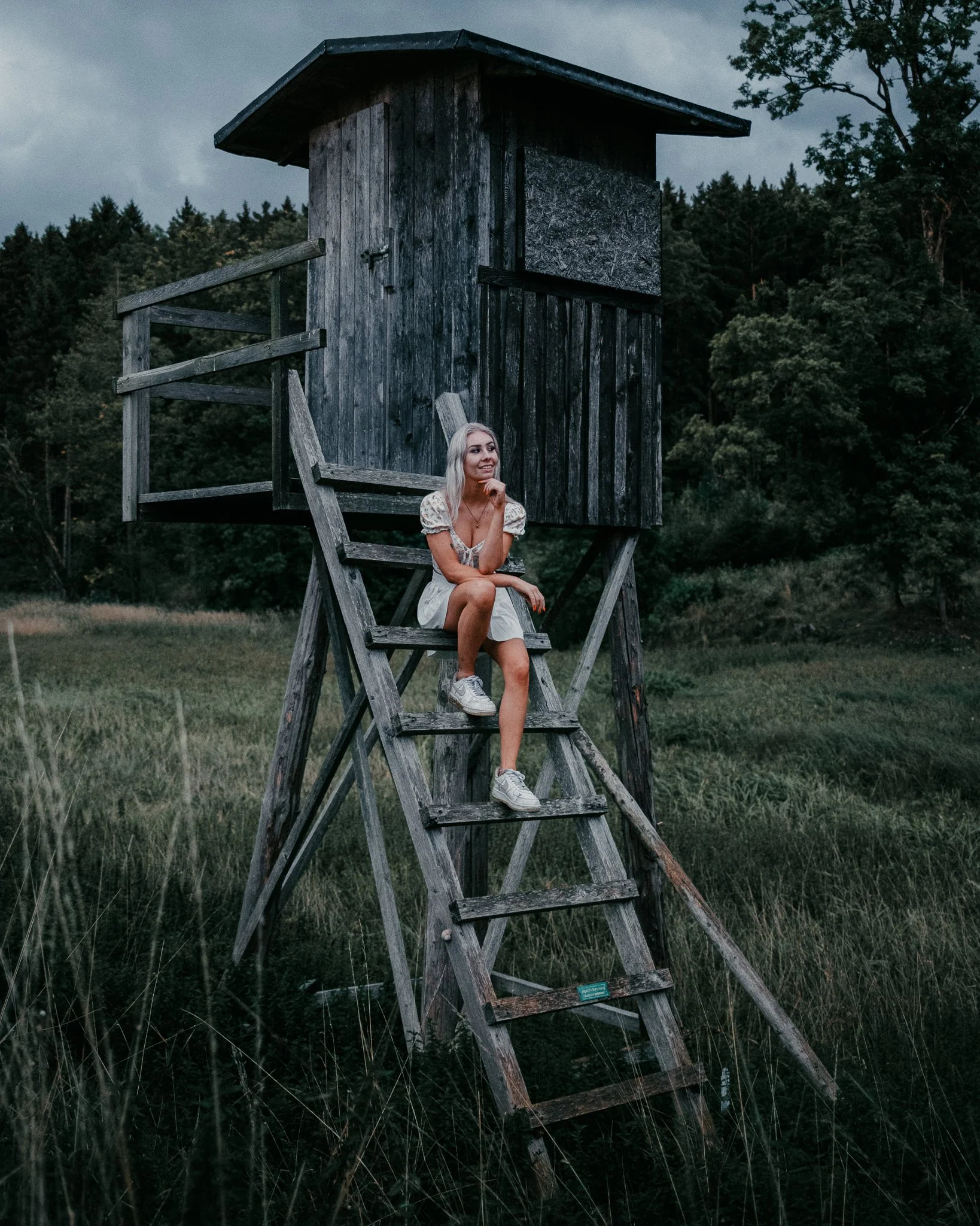 Junge Frau sitzt auf Leiter einer alten, hölzernen Aussichtsplattform in einem Feld mit Bäumen im Hintergrund, bei bewölktem Himmel.