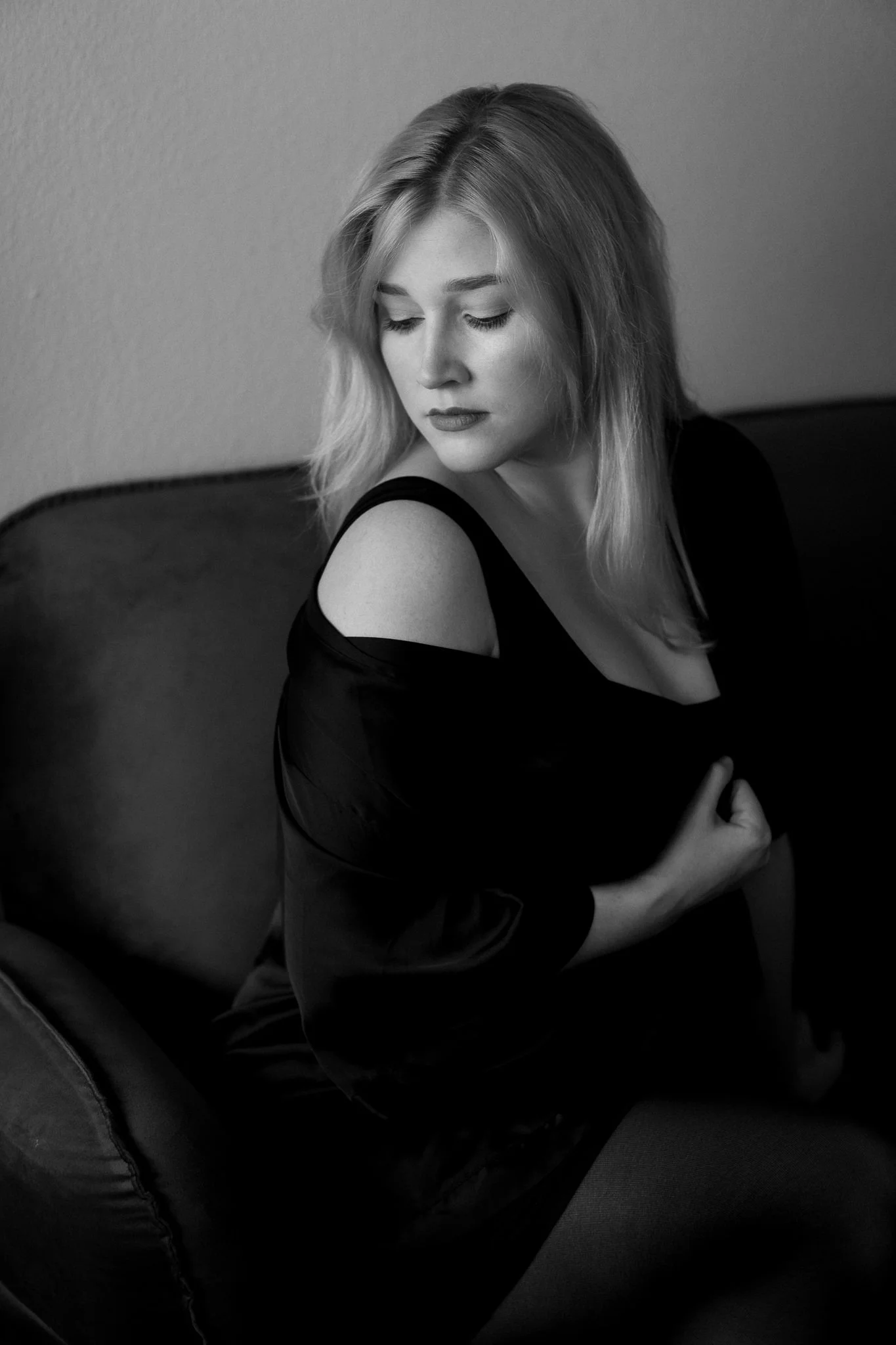 Schwarz-weiß Foto einer jungen Frau mit Schulterlänge Haaren, die auf einem Sofa sitzt, mit Blick nach unten, getragenes dunkles Oberteil mit nacktem Schulterbereich.