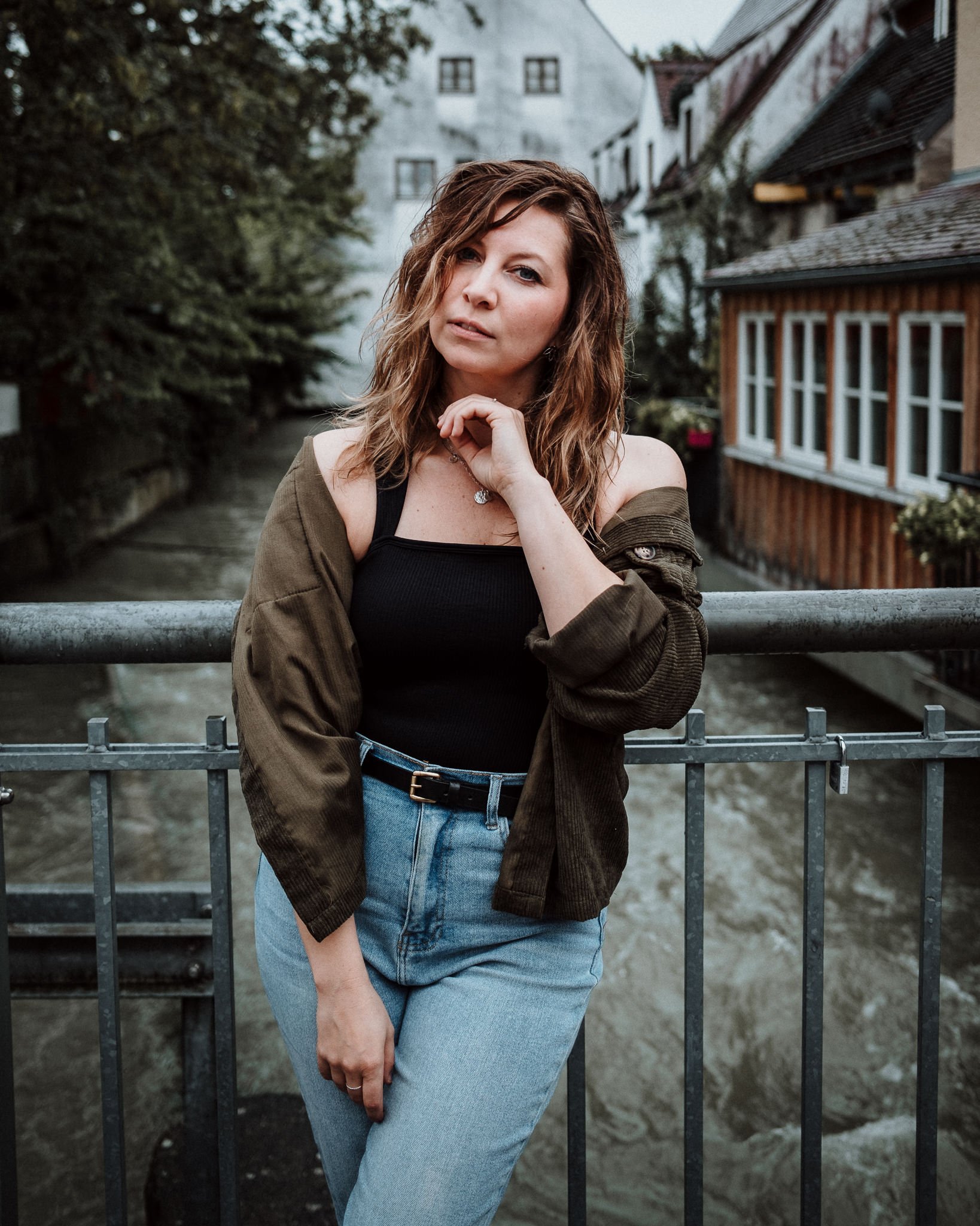 Eine Frau mit lockigem, bräunlichem Haar steht an einem Brückengeländer über einem Fluss, im Hintergrund alte Gebäude und Bäume. Sie trägt ein schwarzes Top, eine dunkelgrüne Jacke und blaue Jeans.