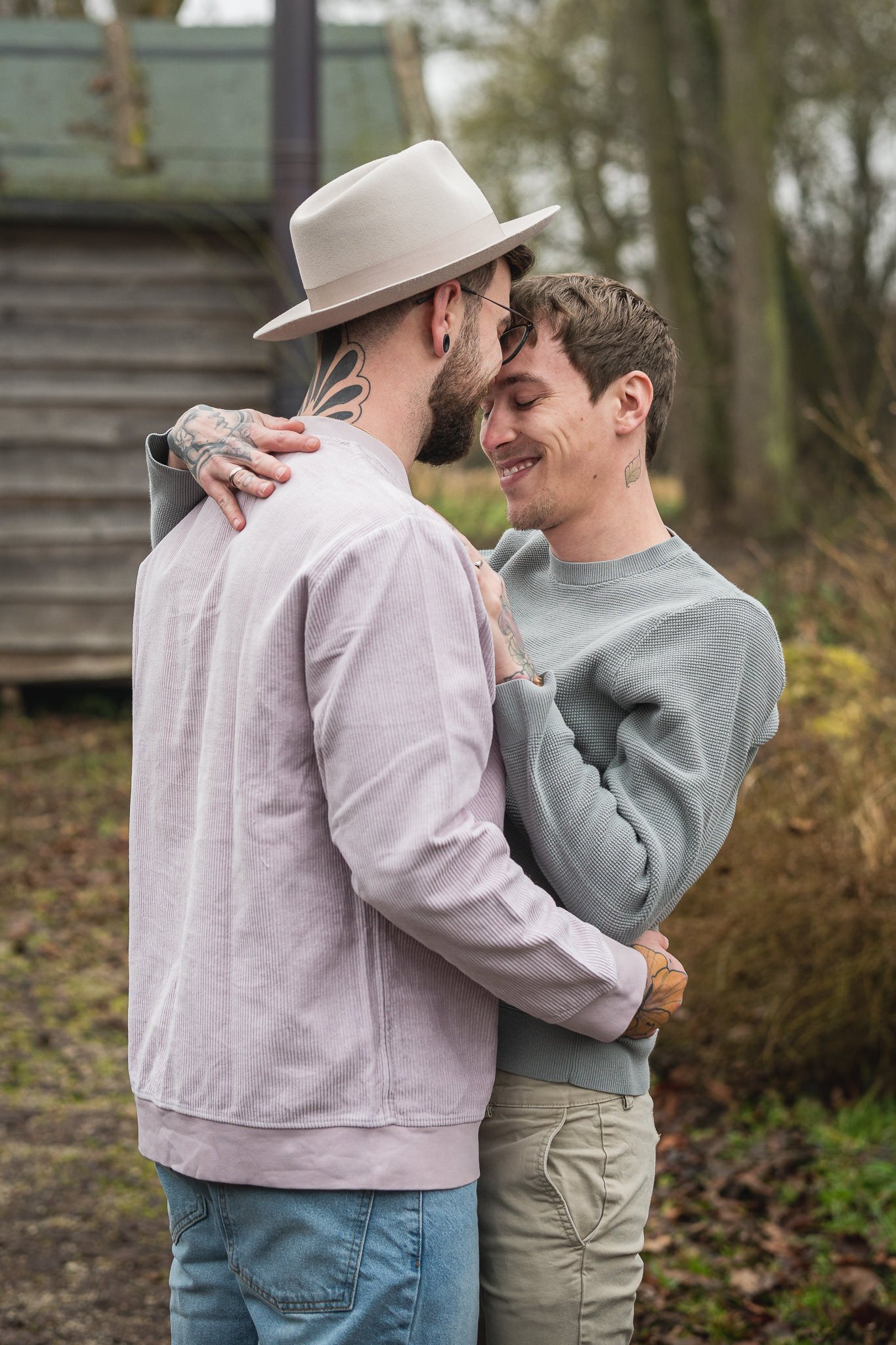 Zwei Männer umarmen sich liebevoll im Freien, lächeln sich an, im Hintergrund Bäume und ein Holzhaus.