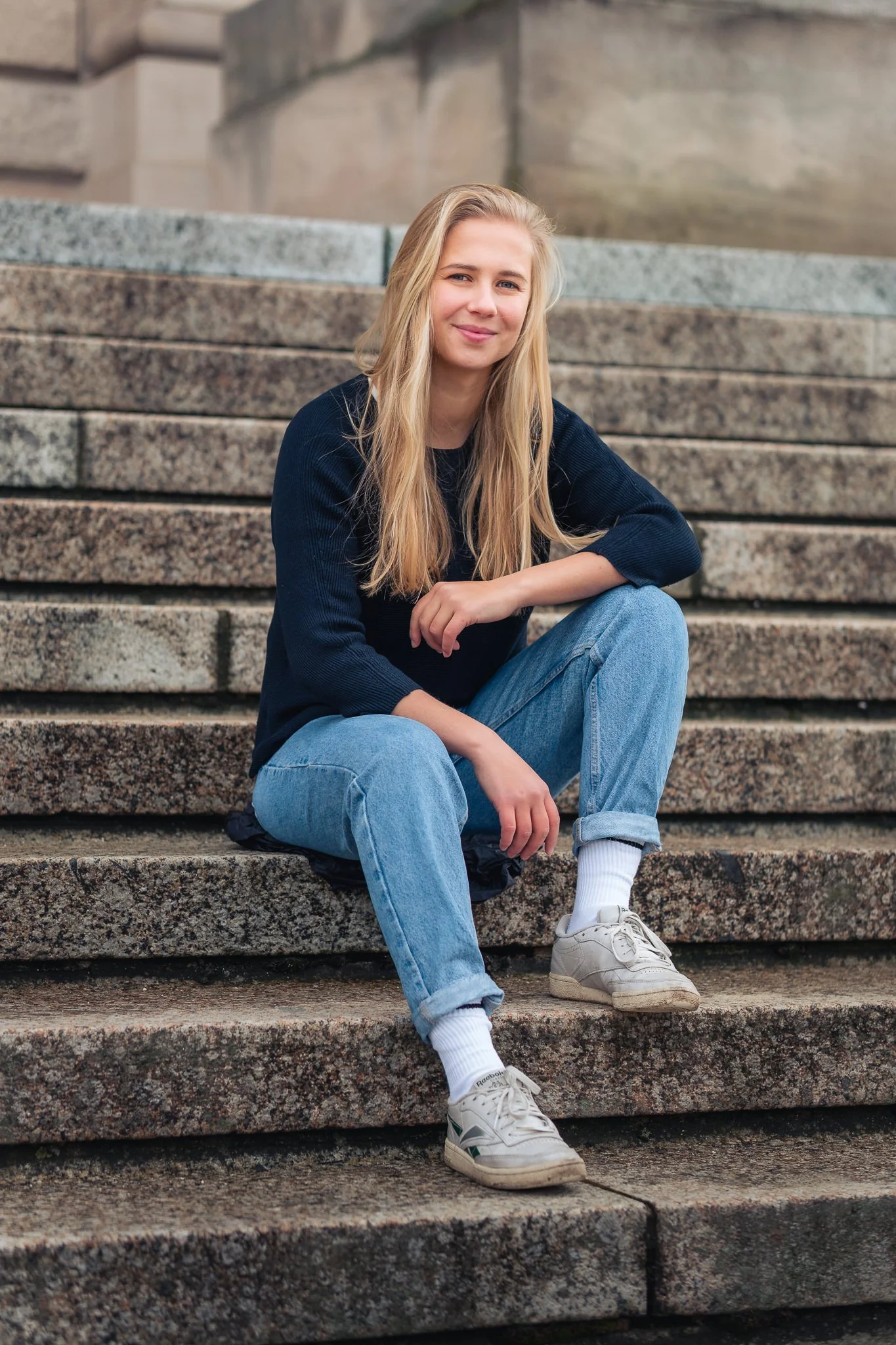 Junge Frau mit blonden Haaren, schwarzem Pullover, blauen Jeans und weißen Turnschuhen sitzt auf Stein-Stufen und lächelt.