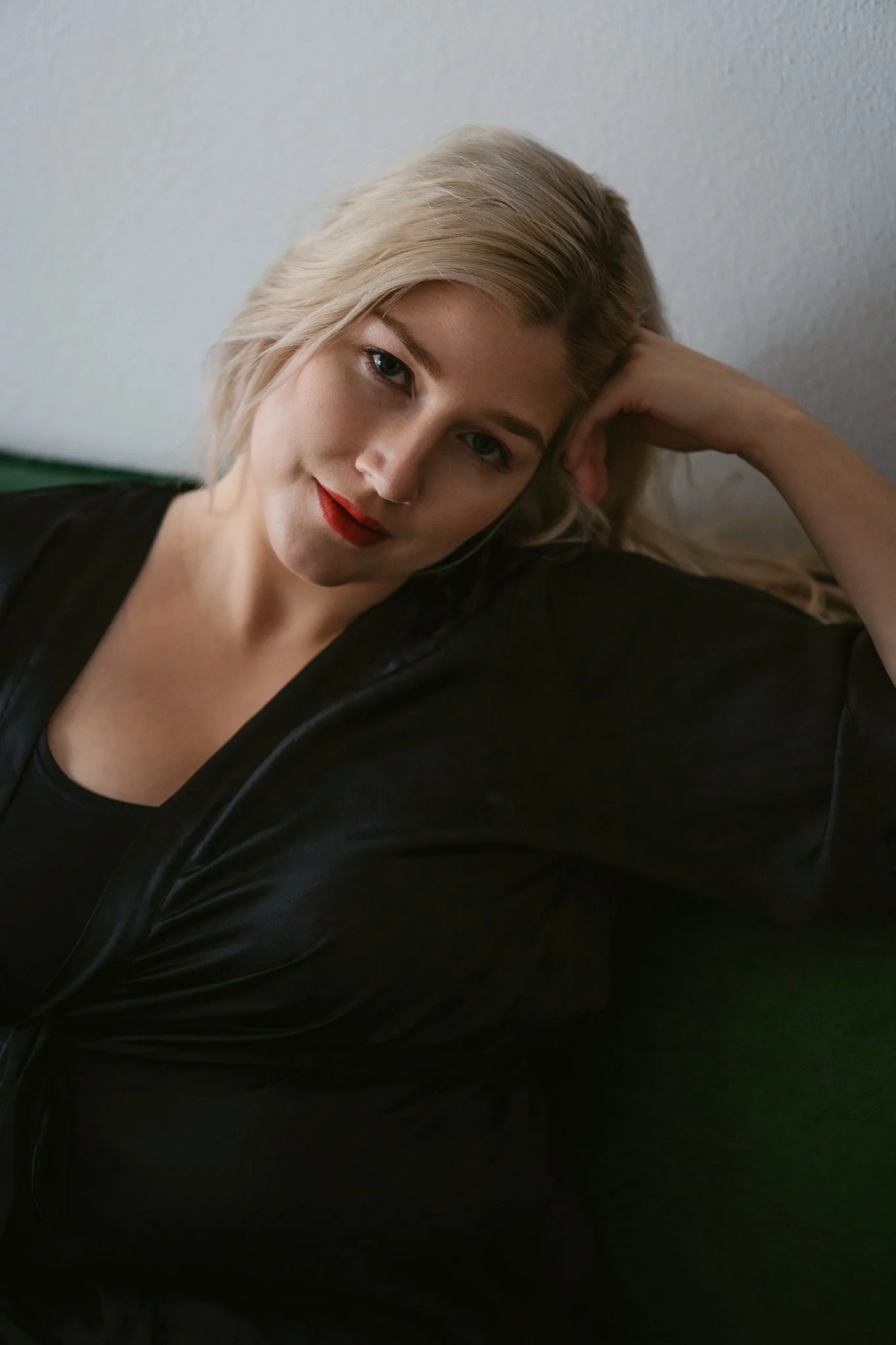 Nahaufnahme einer Frau mit blonden, gewellten Haaren, rotem Lippenstift, schwarzer Kleidung und entspanntem Ausdruck, liegend auf einer Couch oder einem Bett.